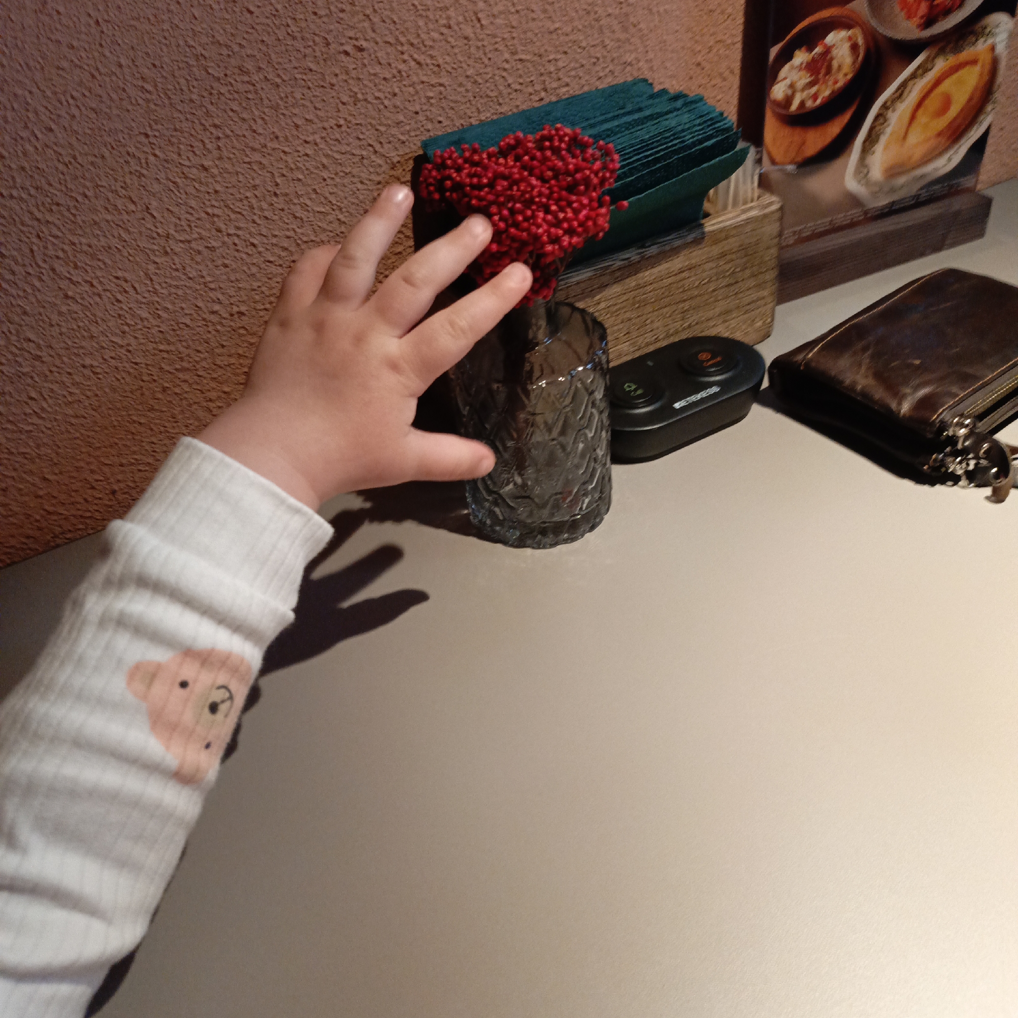 A child's arm and hand are prominently featured on the left, reaching towards a small vase of red berries on a table. The child wears a white long-sleeved top with a light pink teddy bear design on the cuff.
The table setting includes a clear, textured glass vase filled with numerous small, bright red artificial berries, which appear to form a heart shape. Behind the vase is a wooden napkin holder containing neatly folded dark teal napkins. To the right, a black electronic device with "ONCALL" text and two circular buttons (one orange/red, one green) rests on the table. Further right, a brown leather wallet or clutch with a visible zipper and a metal clip attachment is also on the table.
In the background, a menu or poster with blurred images of food, including what appears to be a boat-shaped bread dish (possibly khachapuri) and another bowl of food, is visible. The wall behind the table has a rough, brownish-orange textured finish. The table surface itself is a smooth, light beige or grey. The image depicts a moment of childlike curiosity in what appears to be an indoor restaurant or cafe setting.