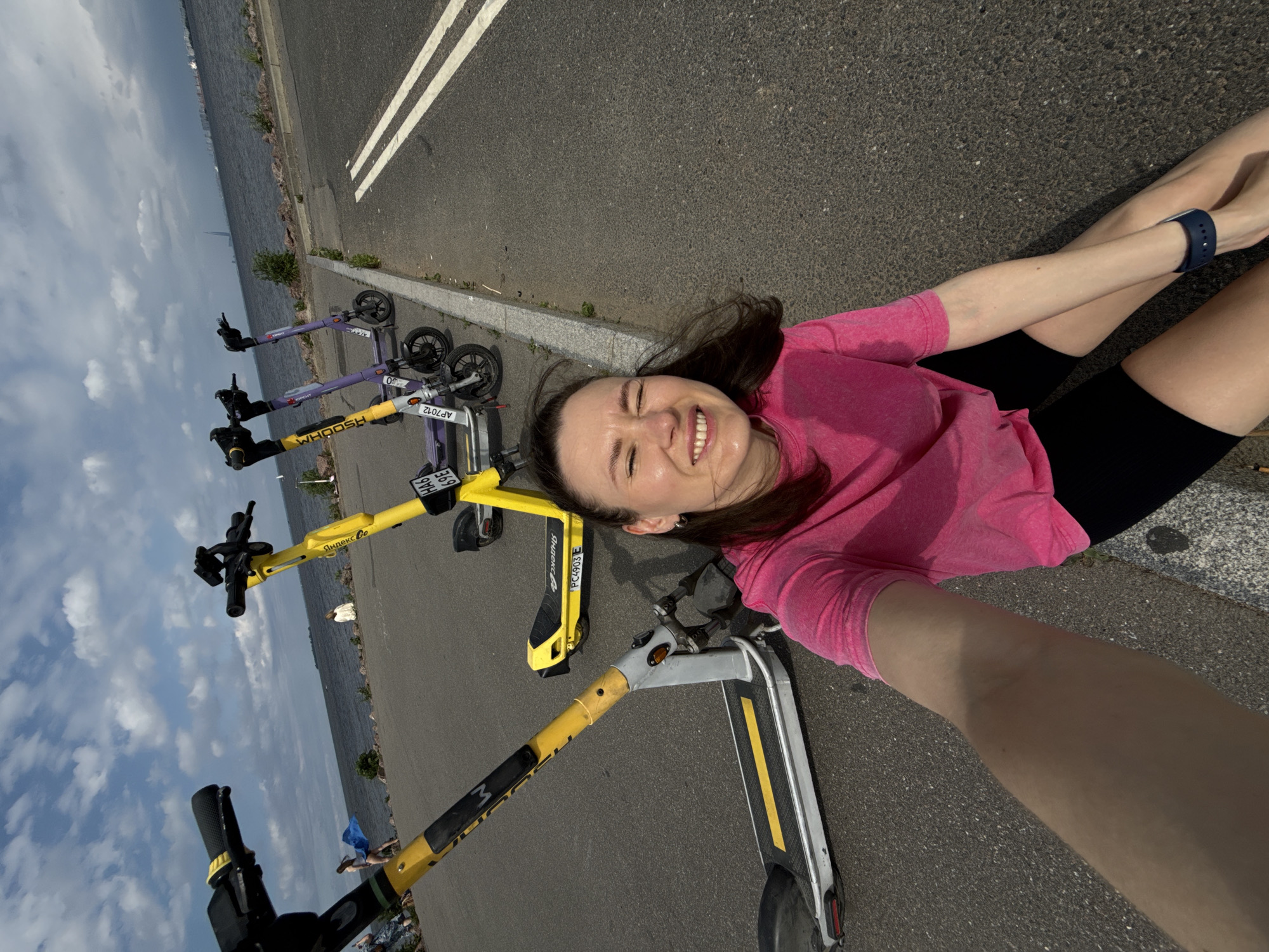 A woman in a pink t-shirt and black shorts is taking a selfie in front of several rental electric scooters parked along a waterfront promenade in Saint Petersburg, Russia. The sky is cloudy, and the water of the Gulf of Finland is visible in the background, with a distant city skyline. The woman is smiling and appears to be outdoors in a park or recreational area.