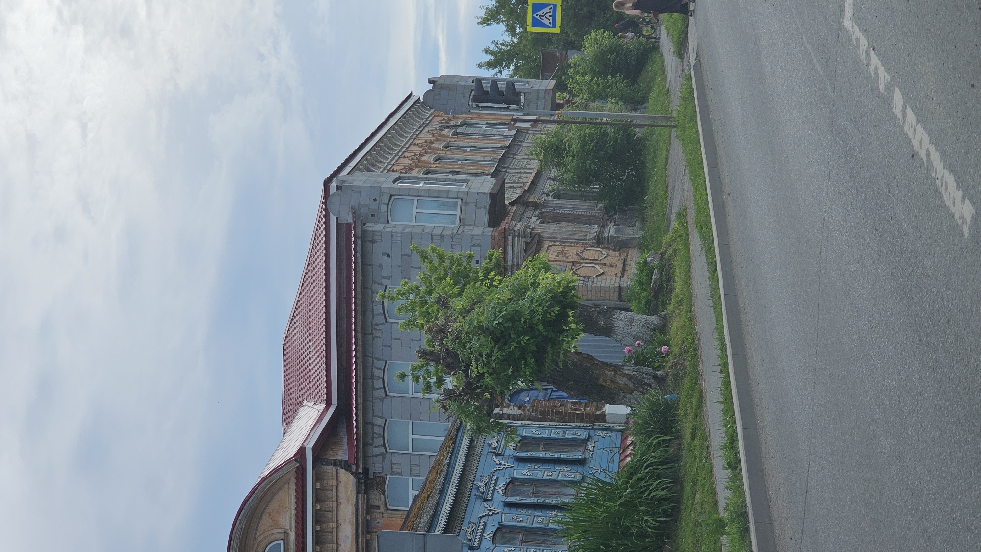 A medium-shot, eye-level, vertical outdoor photograph captures an urban street scene in Kamensk-Uralsky, Russia, on a cloudy day. The foreground features a gray asphalt road with a white dashed line marking a lane. A gray concrete curb separates the road from a grassy verge, where a mature tree with thick, gnarled trunks stands prominently. Pink flowers bloom at the base of the tree. Behind the tree, a section of a blue wooden house with ornate white trim and shutters is visible, followed by a gray, multi-story building constructed of cinder blocks and adorned with decorative elements. The building has a red tiled roof. Further to the right, a traffic light and a pedestrian crossing sign are present. Two people are visible in the background, one on a bicycle and another walking, near the crossing. The sky is overcast with visible clouds.