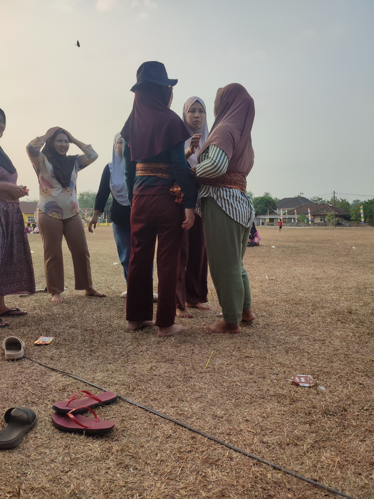 In Nusa Jaya, Indonesia, a group of individuals, primarily women wearing hijabs and modest clothing, are gathered outdoors on a dry, grassy field. One woman in the foreground wears a black fedora and a dark hijab, paired with burgundy pants and a patterned sash. To her right, another woman, also in a hijab, wears a striped shirt and olive-green pants. A third woman, to the left, has her hands on her head, wearing a light-colored patterned top and brown pants. A fourth woman in a long, dark patterned dress stands to the far left. Scattered on the ground are several pairs of flip-flops and some small paper wrappers. In the background, buildings with red tile roofs and a fence are visible, along with a few scattered trees and flags. The sky is overcast and appears hazy. The overall scene suggests a casual outdoor gathering or event.