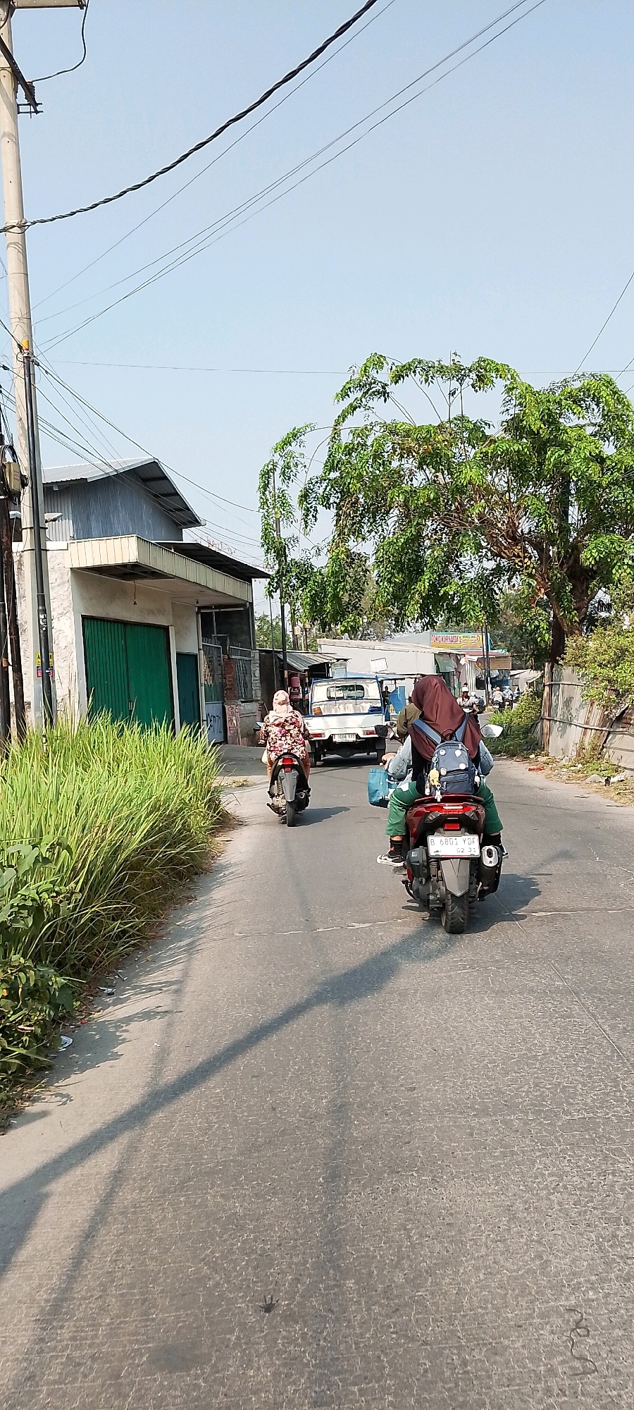 In Babelan, Indonesia, two individuals are riding motorcycles down a paved road under a clear blue sky. The motorcycle in the foreground carries a person wearing a dark red hijab and a blue backpack. The motorcycle behind them has a passenger also wearing a hijab, and the driver is dressed in a floral patterned outfit. On the left side of the road, there is a building with a corrugated metal roof and green metal doors, bordered by tall green grass. A white pickup truck is visible in the background, and a large tree with green foliage stands to the right of the road. Overhead, power lines crisscross the sky, supported by a utility pole. The road is marked with a white line in the center, and shadows from the power lines and a tree are cast across the asphalt.