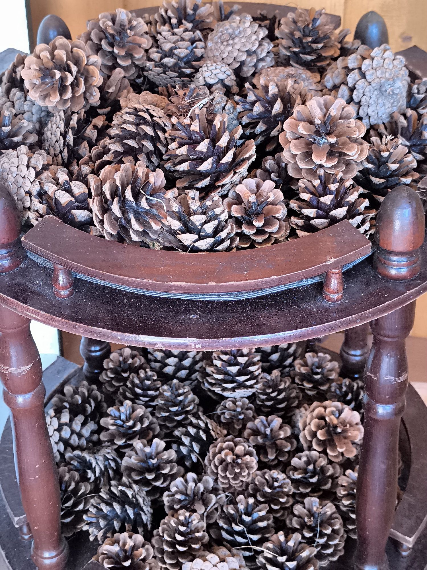 A close-up shot shows a two-tiered wooden stand filled with pinecones. The stand has dark brown wooden posts and two circular shelves. The top shelf is a decorative, curved wooden piece, while the bottom shelf is flat. Both shelves are overflowing with pinecones of varying sizes and shades of brown. The lighting is natural and highlights the textures of the pinecones. The image does not provide any information about the city of Dercekliai, Lithuania, or any people or actions.