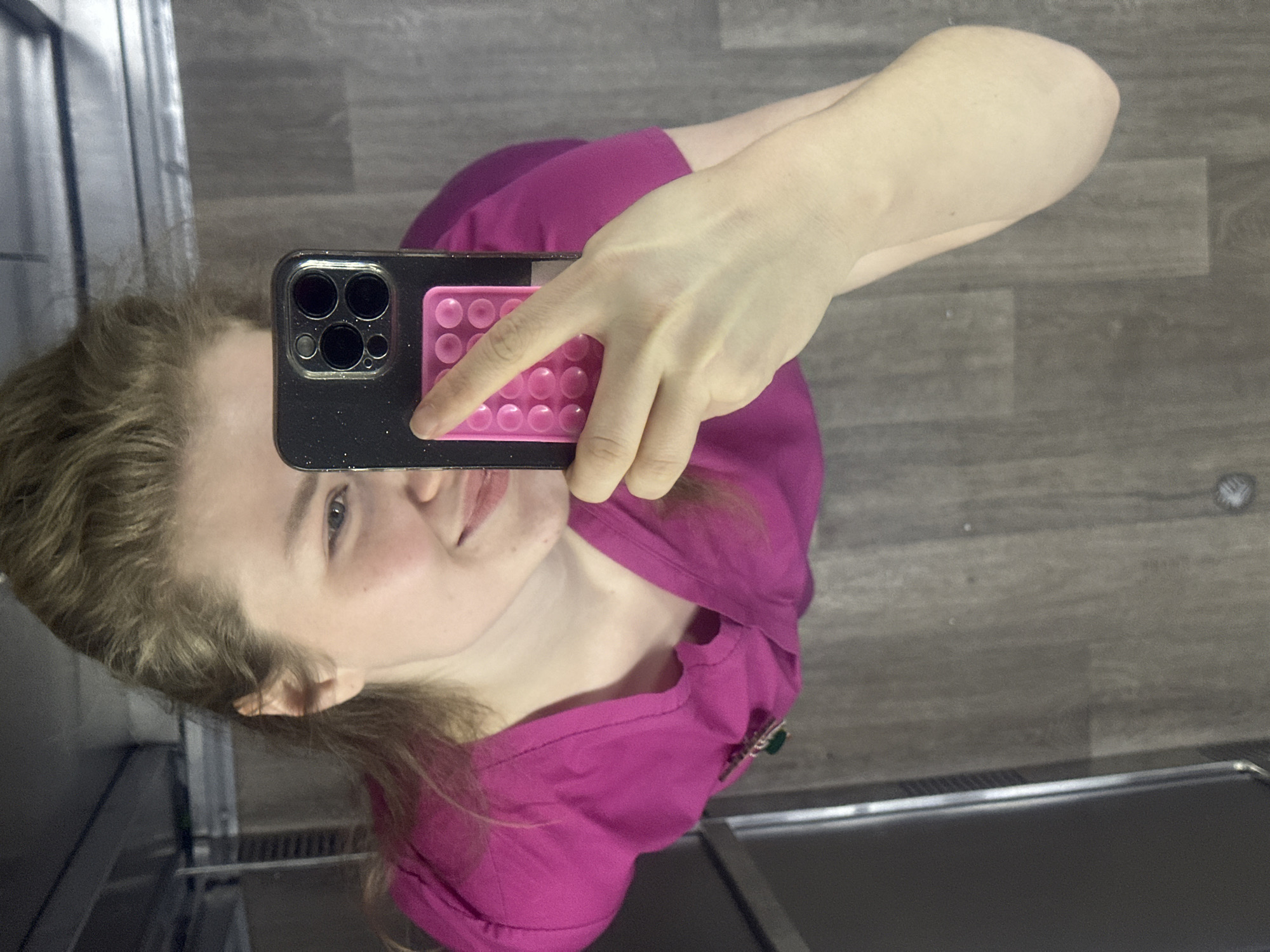The image displays a mirror selfie of a young female, taken indoors in Kazan, Russia. The person, with light brown hair and fair skin, is smiling faintly. She is wearing a fuchsia or magenta-colored top, and a dark green brooch or pendant is visible on her right shoulder.
She holds a dark-colored smartphone, likely an iPhone due to its triple camera lens configuration, in front of her. Attached to the back of the phone is a bright pink, rectangular pop-it style accessory. Her hand is positioned holding the phone, with a finger resting on the pink accessory.
The setting appears to be an interior space, possibly a hallway or elevator, with a gray-brown wooden or wood-laminate floor. Reflections on the left suggest a metallic or mirrored surface. In the bottom right, a dark, possibly upholstered bench or seat with a metallic base is visible. The overall ambiance is modern and indoor.
The image is rotated 90 degrees clockwise, making the person appear on her side. No text is visible in the image.
