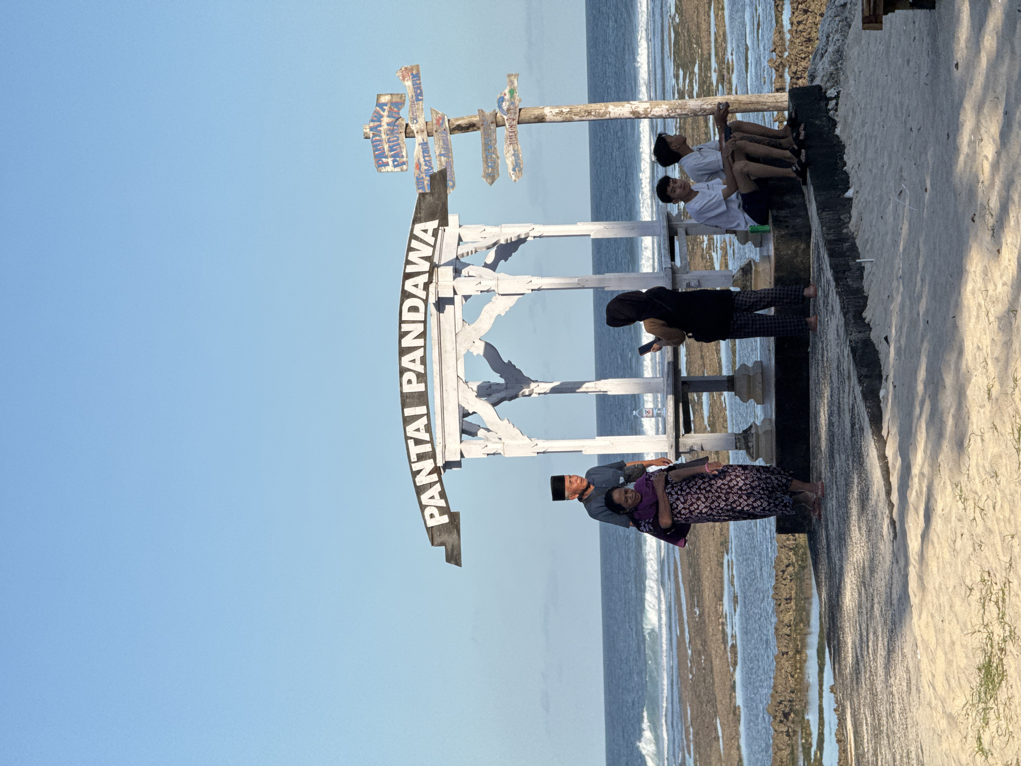 The image captures a group of people posing for a photo at Pantai Pandawa, a beach located in Kuta Selatan, Indonesia. The focal point is a large, white sign reading "PANTAI PANDAWA." The sign is part of a wooden structure with smaller signs pointing in different directions attached to a horizontal beam above. To the right of the sign and structure, several people are sitting on a low concrete wall, facing the ocean. An older couple stands in front of the sign, posing for the photograph. The woman is wearing a dark and light patterned dress, and the man is wearing a gray shirt and a black peci hat. The ocean is visible in the background, and it is dotted with coral. The sky is clear and blue.