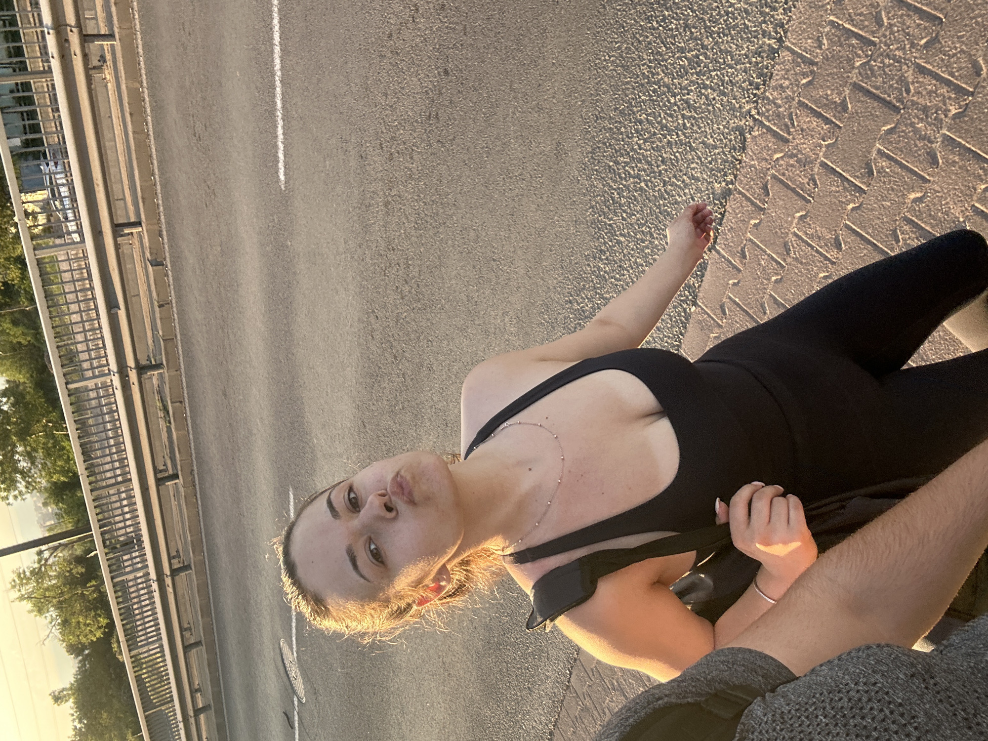 A medium shot photograph depicts a young woman in a black sports bra and leggings, standing on a road with textured concrete on the far right. The road is adjacent to a bridge. The woman has light skin, brown hair, and is making a kissing face towards the camera. Her right arm is extended out. Part of a person's arm is visible on the right side, wearing a grey knit shirt. The location appears to be on a bridge in Sofia, Bulgaria. In the background, there are trees and possibly water visible between the metal bridge structure. A white line is painted on the road. The sun is shining, creating shadows and highlights on the scene.