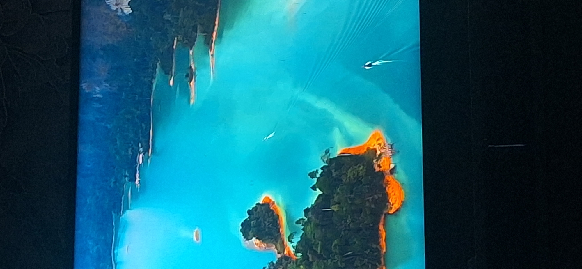 The image displays an aerial view of a vibrant turquoise body of water dotted with lush green islands, some with striking orange shores. The water is clear and reflects the surrounding landscape. Two small boats are visible on the water, leaving wakes behind them. The islands are covered in dense foliage, and one island in the foreground appears to have a small cluster of structures on its orange-colored shore. In the background, the water extends towards a mountainous, forested coastline. The overall scene evokes a sense of natural beauty and tranquility.
The image is not of Bila Tserkva, Ukraine. It depicts a tropical or sub-tropical island landscape.