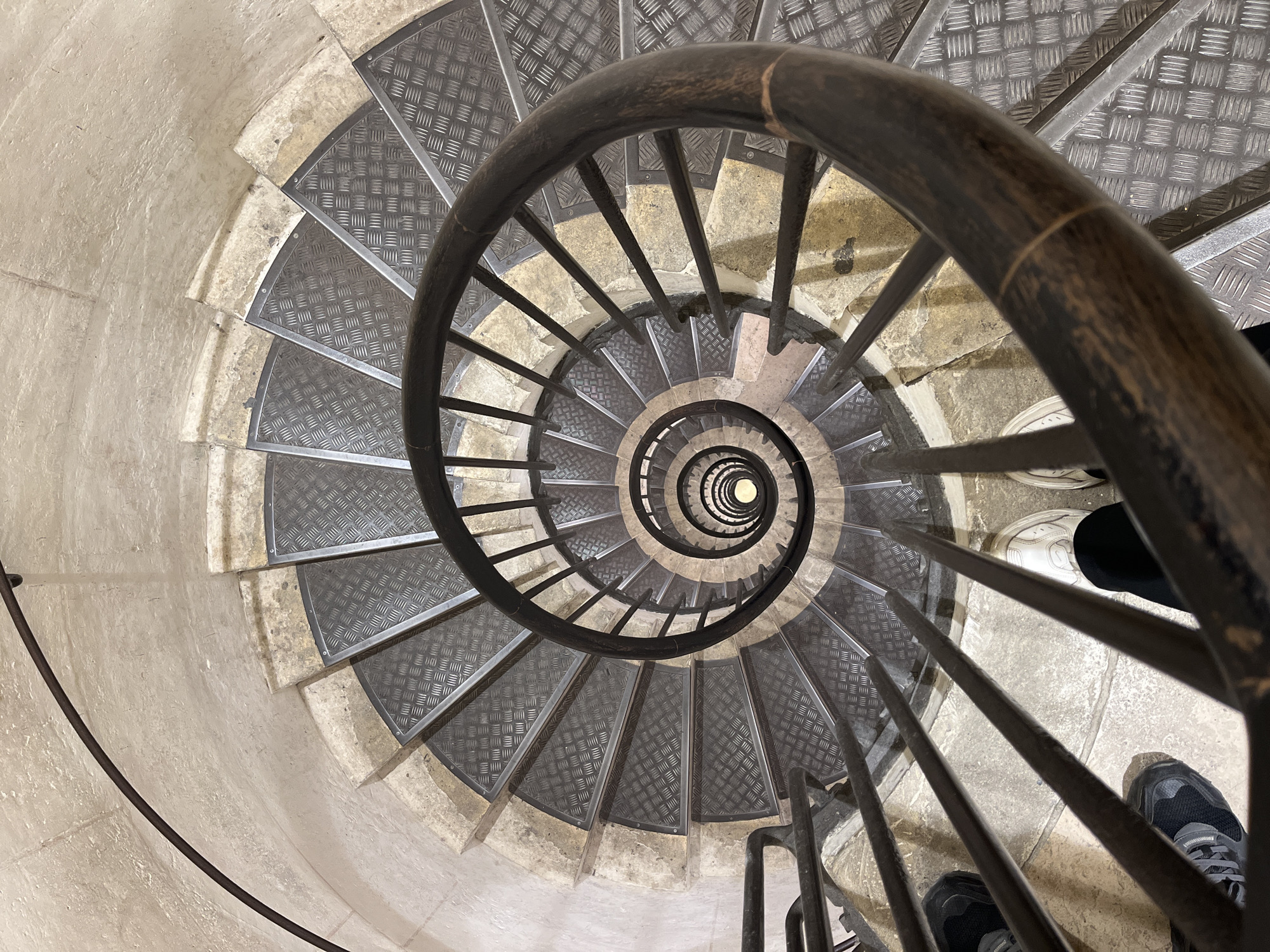 The image captures a top-down view of a spiral staircase, emphasizing its descent into a deep well. The staircase is constructed with light-colored, aged stone or concrete steps, topped with dark gray metal checker plate (diamond plate) treads. Each step is trapezoidal, forming a continuous spiral. A dark, possibly black or very dark brown, metal handrail follows the curve of the stairs, supported by thin vertical metal bars that extend from the railing to the steps below. The handrail shows signs of wear, with lighter brown scuff marks visible.
The perspective is directly overhead, looking down into the center of the spiral, which narrows significantly towards the bottom, where a small, bright circular opening is visible, suggesting a light source or an opening to another level. On the upper right side of the image, the feet of a person are visible, wearing a white sneaker on the left foot and a dark grey sneaker on the right foot, implying the observer is standing on the stairs or at the top looking down. The lighting is diffused, illuminating the steps and the well of the staircase evenly, with some natural shadows accentuating the depth. There is no visible text. The location context given is Paris, France, although no specific landmarks are identifiable in the image itself.
