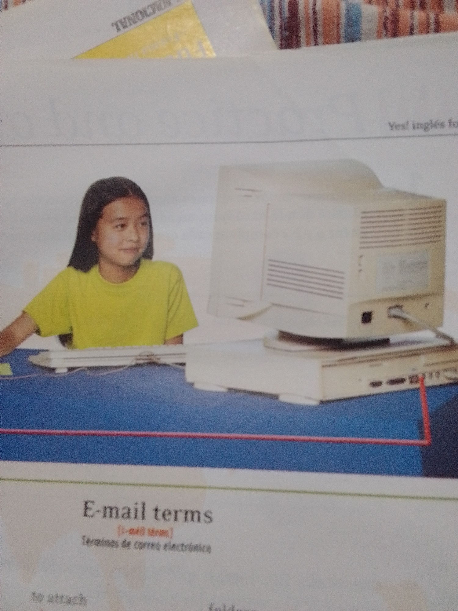 The image shows a young girl with dark hair, wearing a yellow t-shirt, sitting at a table in front of a vintage computer. The computer consists of a beige monitor and a beige tower. A keyboard is in front of the girl, and a red line is drawn across the image, framing the computer setup. The background is a plain white wall with some blurry, colorful stripes at the top. Below the computer and the girl, text is visible, including "E-mail terms" and "Términos de correo electrónico." The scene appears to be in a classroom or a study area. There is no indication of the city of La Tagua, Venezuela, in the provided image.