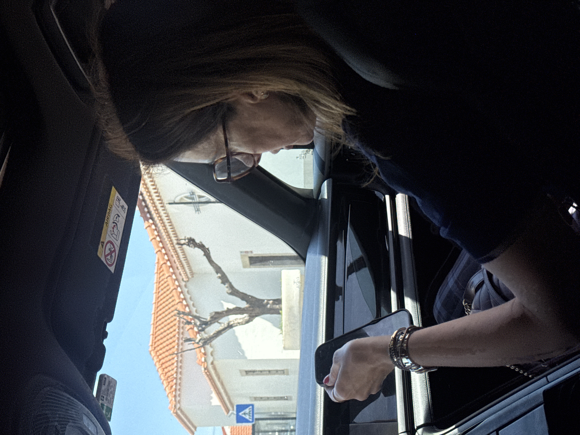 A high-angle shot captures a person, likely a woman, leaning out of a vehicle's open window. She has brown hair, wears glasses, and an earring is visible on her right ear. Her right arm, adorned with multiple bracelets and a watch, extends out of the window, holding a smartphone in her hand. She appears to be looking down and interacting with the phone or something outside the vehicle.
The interior of the vehicle shows parts of a dark fabric seat, the door frame, and a sun visor with an "AIRBAG" warning sticker and related symbols.
Through the open window, a sunny outdoor scene is visible under a clear blue sky, suggesting it is midday or early afternoon. A building with a traditional orange tiled roof and white walls occupies the background. A large, bare tree branch extends across the view in front of the building. Near the bottom left, a blue triangular road sign with a pedestrian crossing symbol is partially visible. The bright light casts distinct shadows. The setting appears to be a street in an urban or semi-urban area, consistent with Torres Vedras, Portugal.