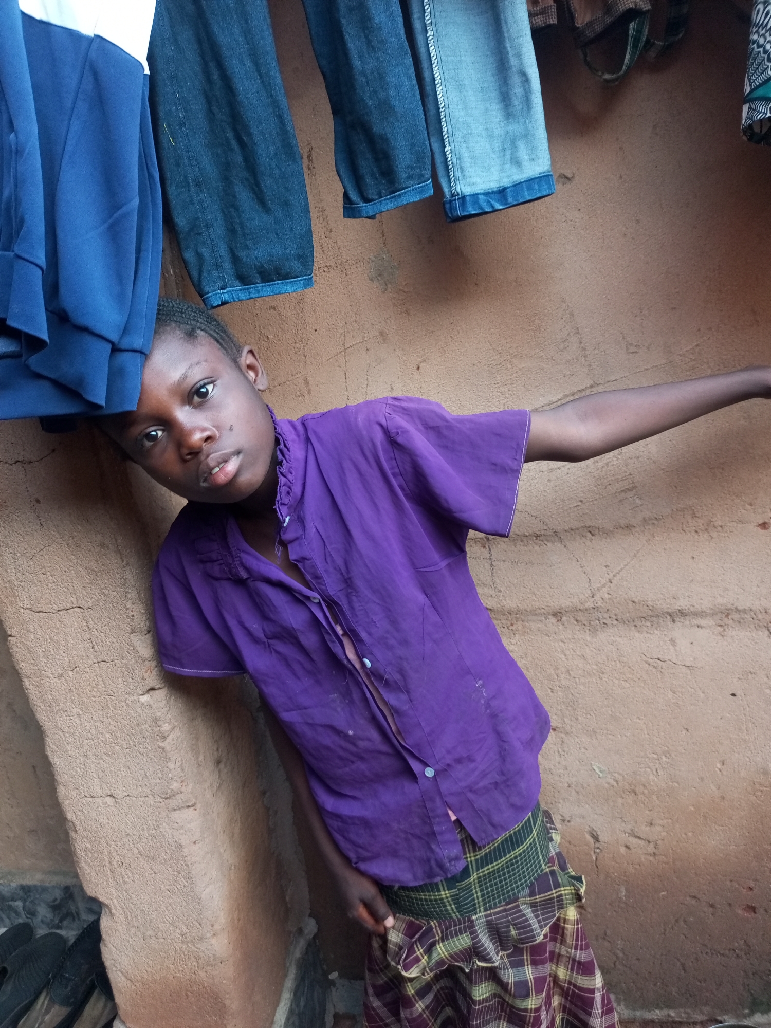 In Tempure, Nigeria, a young Black individual is captured in a medium shot, leaning against a rough, light brown wall. The individual wears a vibrant purple, collared shirt with short sleeves and a plaid skirt featuring green, yellow, and maroon checks. Their left arm extends horizontally across the frame, while their right arm is bent at the elbow, resting by their side. The background is dominated by the textured wall and clothing items suspended from above, including what appears to be blue fabric and denim jeans. In the lower left corner, a pair of black sandals rests on the floor.