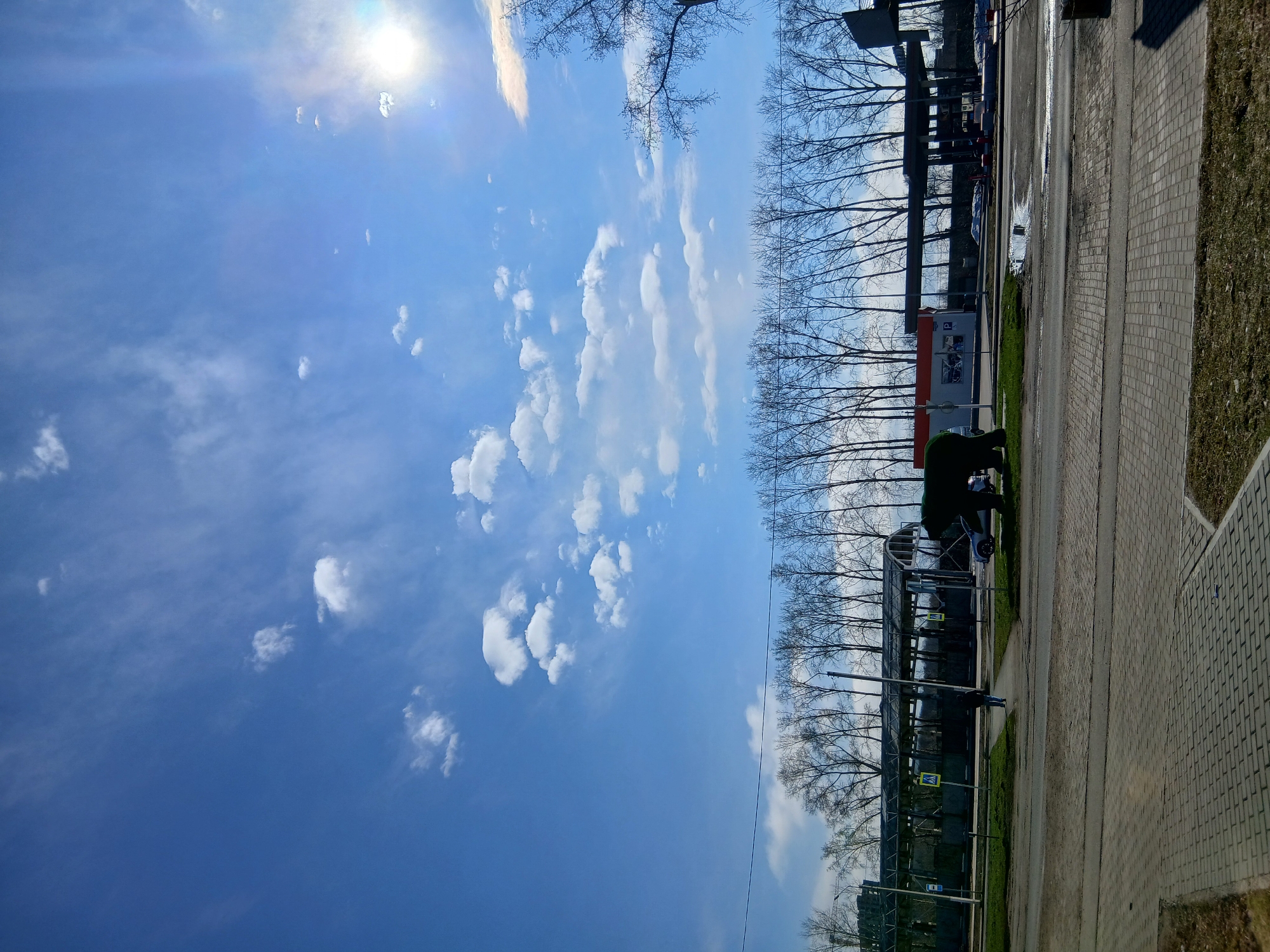 The image shows an outdoor scene with a clear blue sky with scattered white clouds and a bright sun. On the right side of the frame, there is a paved sidewalk made of rectangular blocks. Adjacent to the sidewalk is a patch of green grass and a row of trees without leaves.
A bus stop is visible, with a metal structure and signage. There are some people waiting at the bus stop. A building and additional trees can be seen behind the bus stop. The location is in the city of Unknown, Unknown.