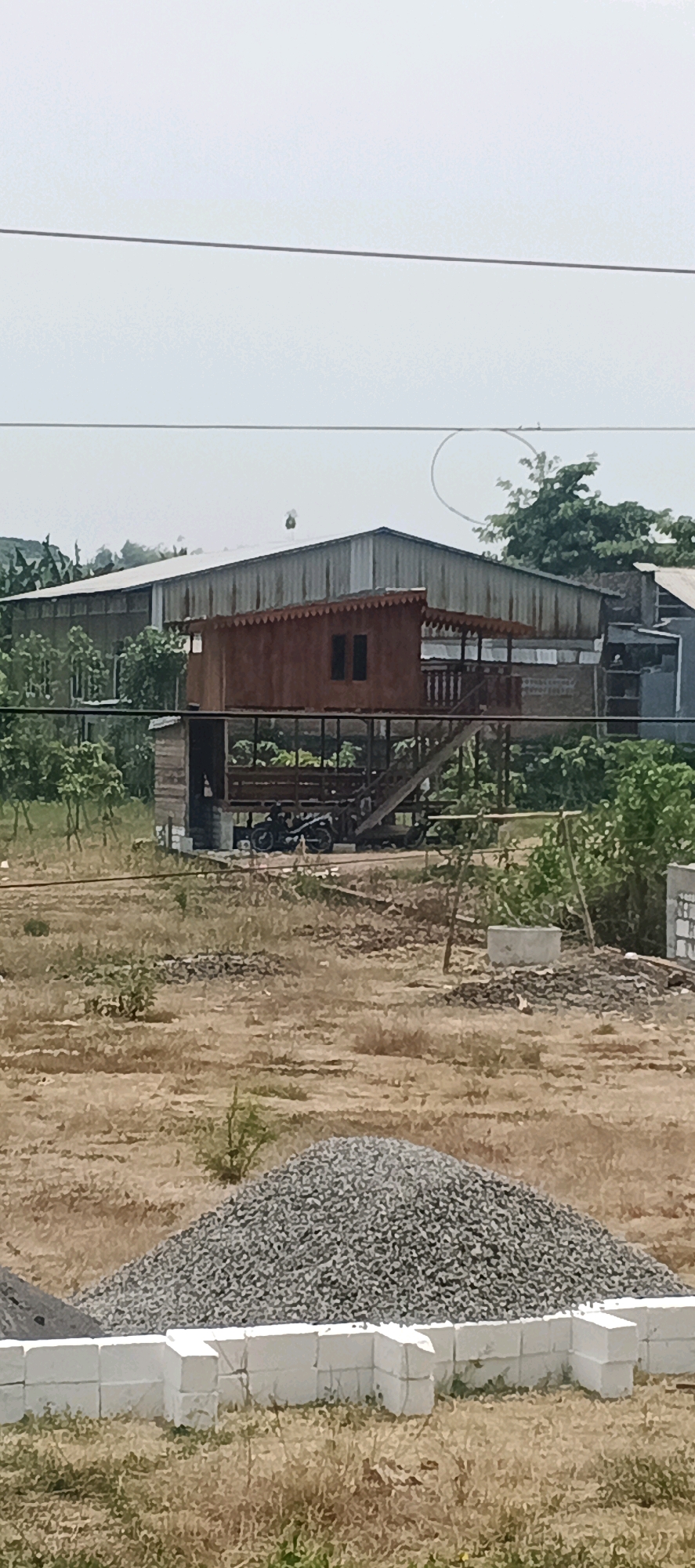 The image depicts a rural scene in Laren, Indonesia, likely a construction or renovation area. In the foreground, a pile of grey gravel sits behind a low wall constructed from white concrete blocks. The middle ground features a raised wooden structure, resembling a small house or platform, with a brown, corrugated roof and dark windows. Stairs lead up to this structure, and motorcycles are visible parked underneath it. Surrounding this structure is dry, brown vegetation. In the background, a large, corrugated metal industrial building dominates the skyline, with some greenery and smaller buildings adjacent to it. The sky is bright and overcast.
