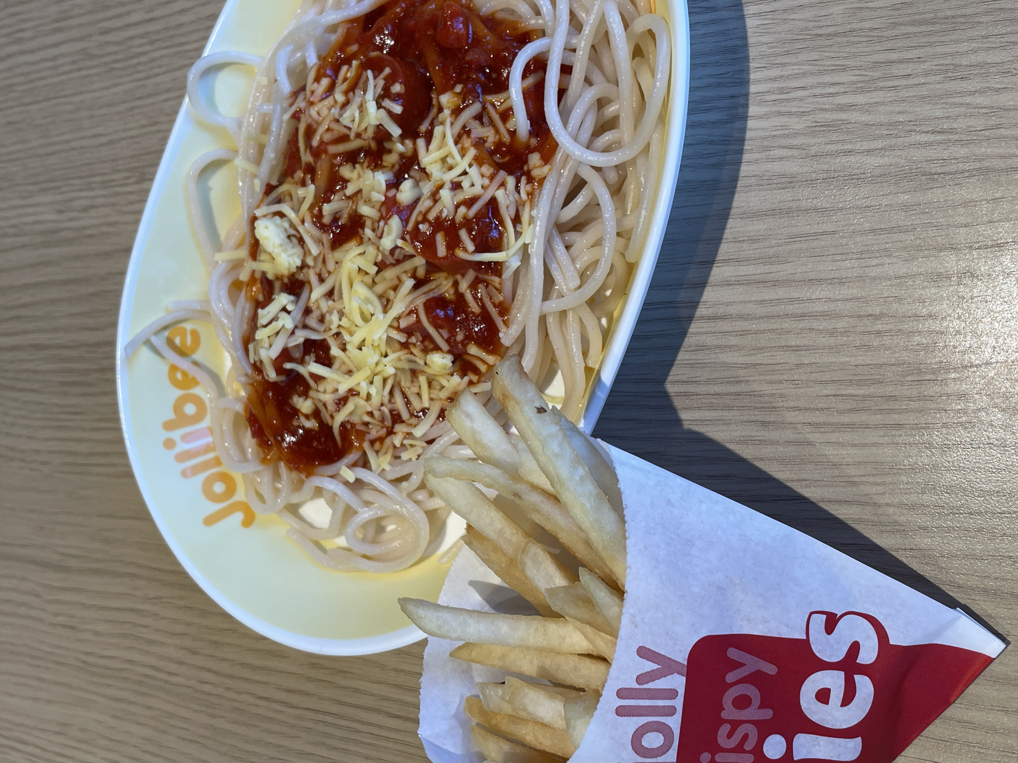 This media file shows a close-up, overhead shot of Jollibee spaghetti and french fries, likely a meal. The spaghetti is served on a yellow Jollibee branded plate, topped with a hearty portion of red sauce and grated cheese. The french fries are presented in a white paper bag with red accents, featuring the Jollibee logo and text. The food is placed on a light-colored, wood-grain surface, suggesting a table. The lighting indicates it's daytime, possibly indoors with natural light contributing to the scene. The image is well-lit and the focus is sharp on the food, highlighting the textures of the spaghetti, sauce, cheese, and fries. There is a shadow cast on the table, possibly from the food items or an unseen object, suggesting the light source is from the right. The overall impression is that of a casual meal. Visible text includes "Jollibee" on the plate and "Jolly Crispy Fries" on the paper bag. The context suggests this is likely in Davao City, Philippines, a common location for Jollibee establishments.