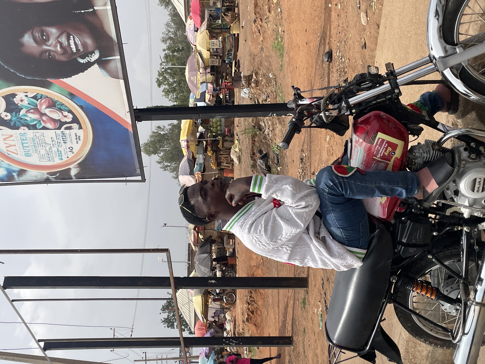 A person is sitting on a motorcycle in what appears to be an outdoor market setting in Bar Jirgi Summa, Nigeria. The market is visible in the background, featuring various stalls and vendors under umbrellas. A large advertisement billboard for "ZYN Refined Shea Butter" with a smiling woman's face is prominently displayed in the left side of the frame. The motorcycle is in the foreground. The person is dressed casually and appears to be relaxing or waiting. The ground is reddish-brown dirt.