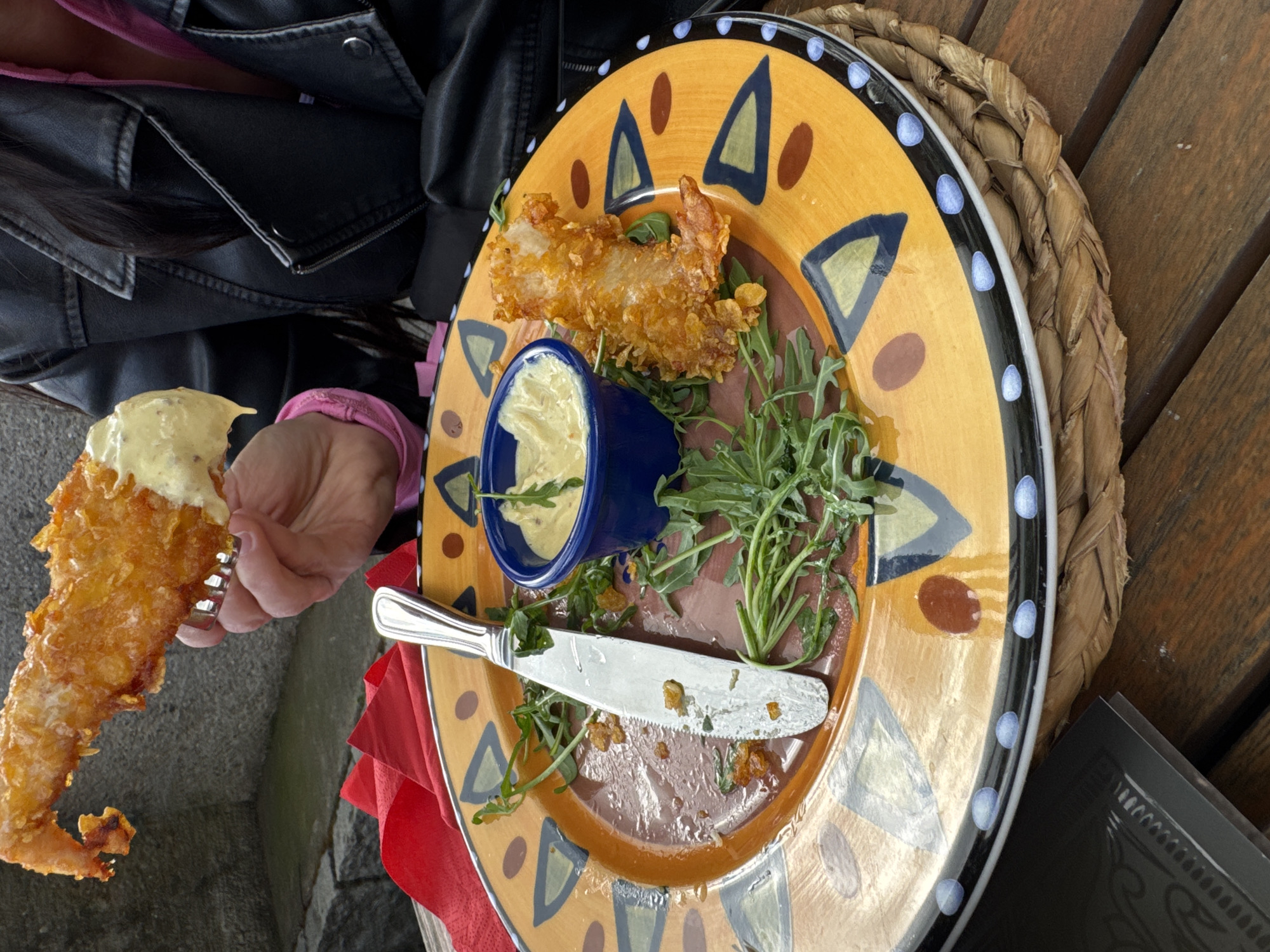 The image shows a partially eaten plate of food, likely fried shrimp or prawns, with a side of sauce in a blue container and some greens. A knife is resting on the plate. A hand holds a piece of the fried shrimp, dipped in sauce. The plate is yellow with a patterned rim and sits on a woven placemat or basket. A portion of a black leather jacket and pink shirt are visible in the upper left corner of the frame. Based on visual analysis, the image appears to be taken at a restaurant in the city of Backnang, Germany.