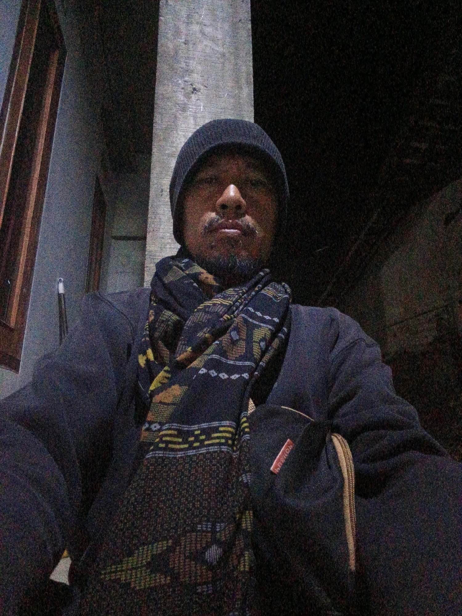 A medium-shot selfie shows a man with a beard and a beanie, wearing a scarf and a dark jacket. He is standing outside at night, with a blurry background that appears to be a wall and a window. The man is looking directly at the camera. The image is grainy, suggesting it was taken in low light conditions. The man is positioned in front of a pillar and a building structure.