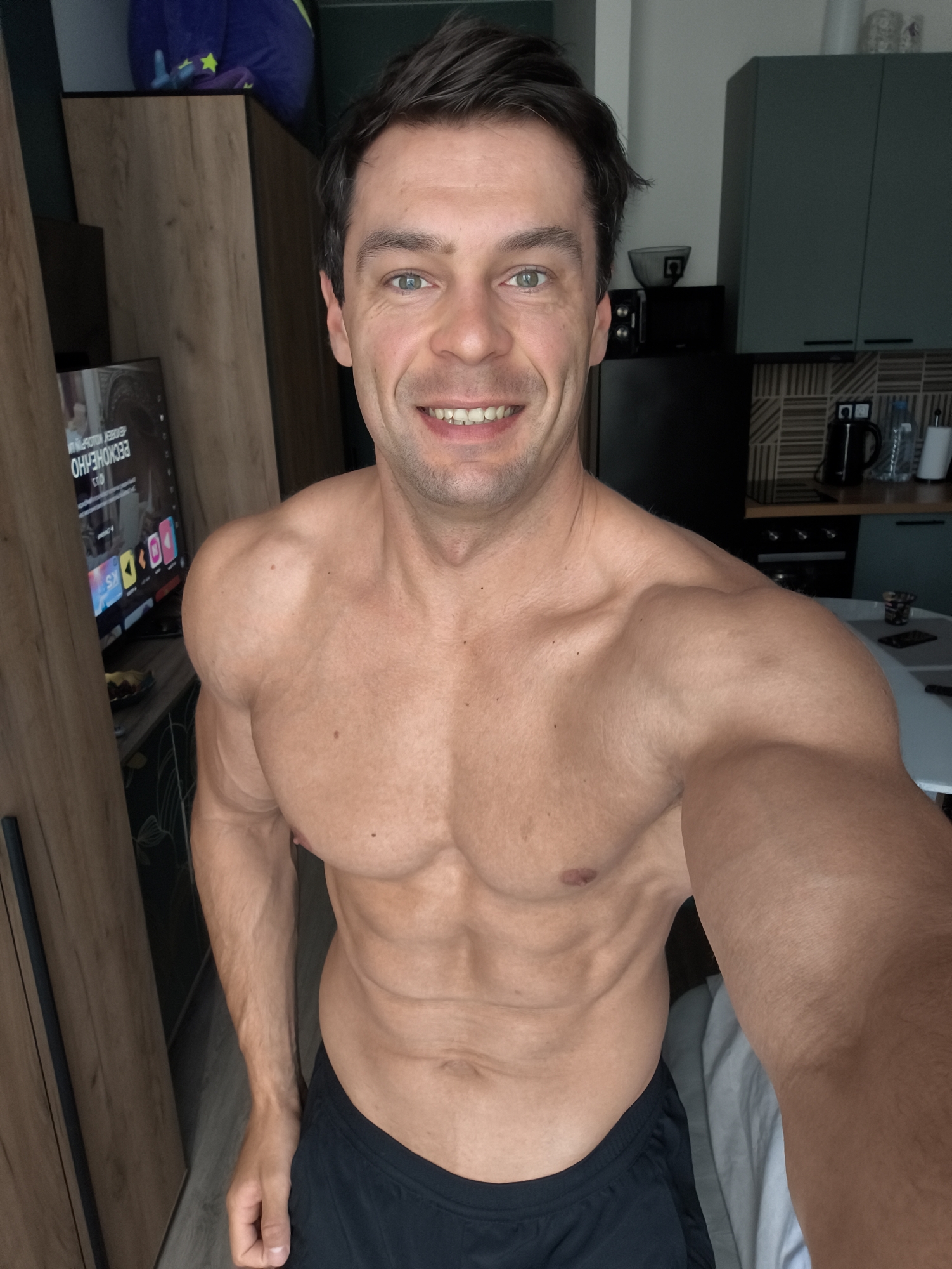 A lean and muscular man, appearing to be in his 30s or early 40s, poses for a shirtless selfie indoors. He has short, dark hair and striking green or hazel eyes, smiling broadly at the camera. His athletic physique displays prominent abdominal muscles, well-defined deltoids, and toned arms. He is wearing black athletic shorts.
The setting is an apartment, featuring an open-plan layout that includes a living area and a kitchen. To the man's left, a wooden wardrobe stands beside a television screen. The TV displays a streaming interface, with the Cyrillic word "БЕСКОНЕЧНО" (BESKONECHNO), meaning "infinitely" or "endlessly," clearly visible. A blue and purple object, possibly a toy or pillow, rests on top of the wardrobe.
To the man's right, a modern kitchen is equipped with dark green overhead cabinets, a black refrigerator, and a microwave. The counter holds a black electric kettle, a water bottle, and a white cylindrical object, against a backsplash with a geometric tile pattern. A light-colored table is partially visible. The bright natural lighting suggests it is daytime. The man's expression conveys happiness and confidence. The Cyrillic text on the TV, combined with the contextual information, suggests the location is Moscow, Russia.