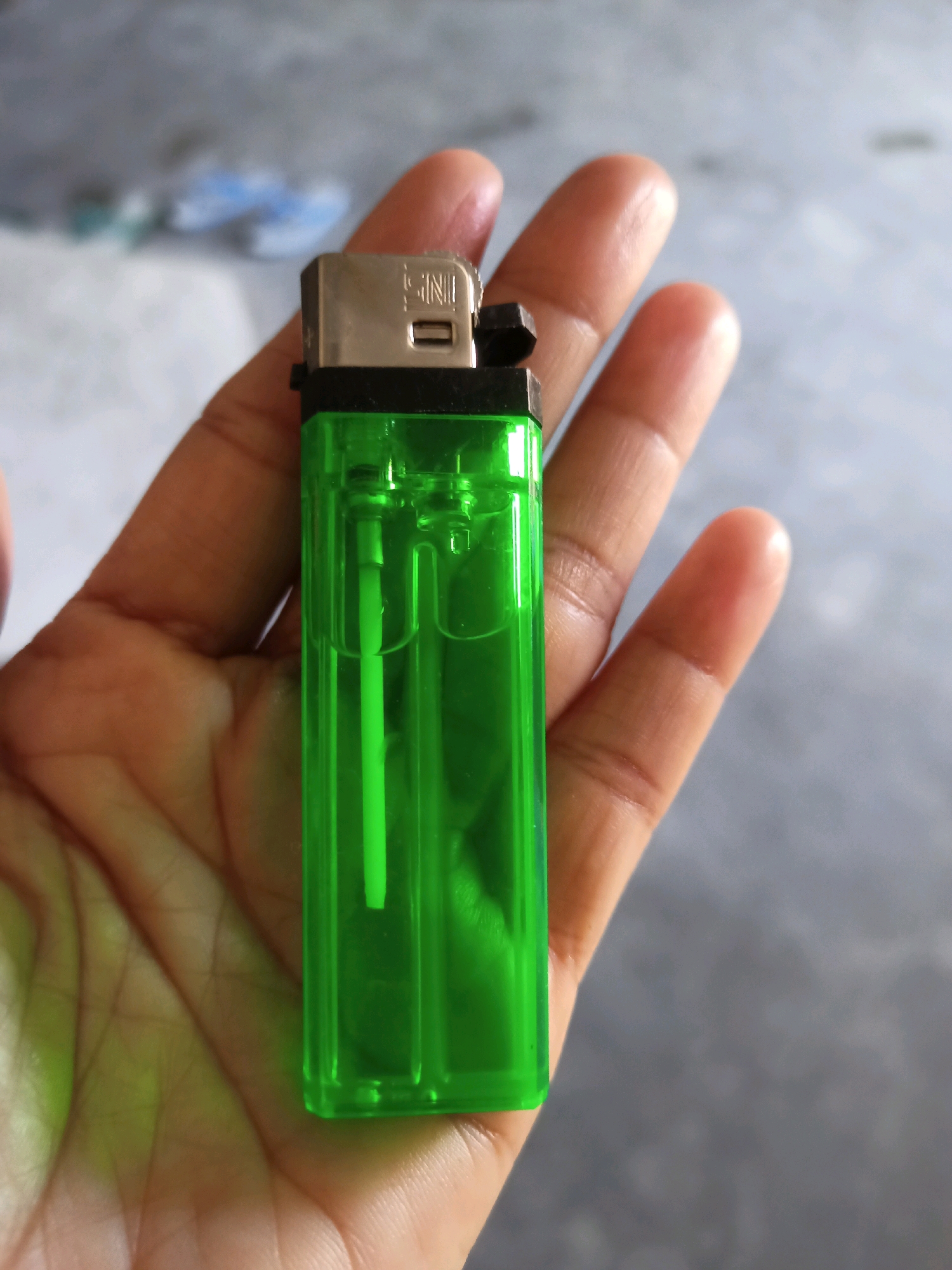 A hand holds a bright green disposable lighter. The lighter is made of clear plastic and contains clear liquid fuel. The metal top of the lighter has the inscription "SNI" stamped onto it. The hand is cupped and the lighter rests in the palm, held by the thumb and forefinger. The background is blurred and appears to be an outdoor setting with a gray concrete surface. The image was taken in Karangtalun, Indonesia.