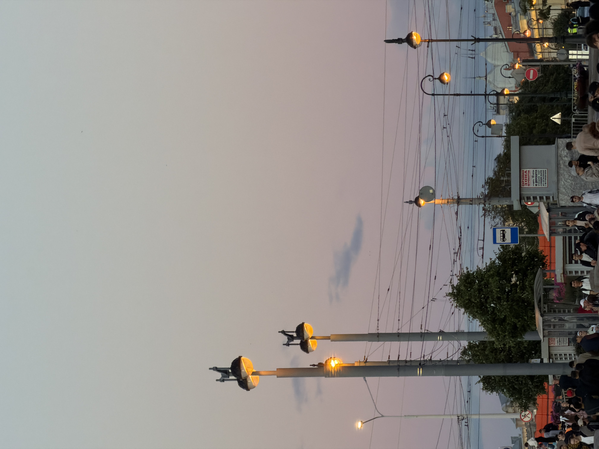 A street scene in St. Petersburg, Russia, captures the twilight hour with a soft, pastel-colored sky transitioning from pale blue to pink. Several tall lampposts with ornate designs and glowing yellow lights are prominent, some featuring multiple lamps. Overhead, a complex network of electrical wires crisscrosses the sky, suggesting a city with active public transportation.
In the foreground, a crowd of people is gathered. They appear to be waiting, possibly at a bus stop, as indicated by a blue bus stop sign visible in the mid-ground. Some individuals are interacting, while others stand individually. The architecture in the background includes buildings with classical elements and domes, characteristic of St. Petersburg. Trees are interspersed throughout the scene, adding a touch of greenery to the urban landscape. The overall atmosphere is one of urban activity at the close of day, with the artificial lighting beginning to dominate the fading natural light.