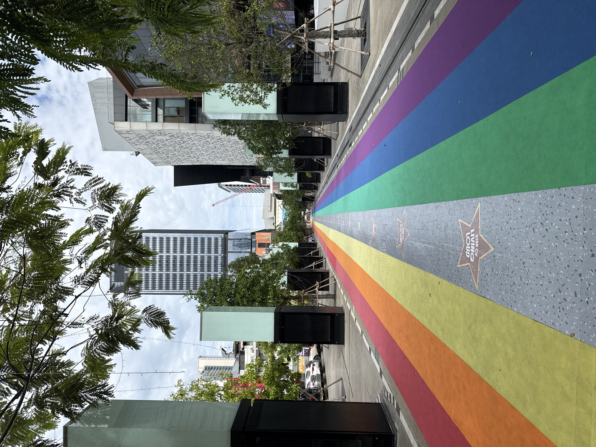 The image depicts a street in Bangkok, Thailand, decorated with a rainbow-colored carpet that extends down the middle of the road. The street is flanked by modern buildings, trees, and tall, illuminated pillars. Several cars are parked on the side of the street, and a few people are visible in the distance, walking along the rainbow path. The sky is overcast, and the overall atmosphere suggests a public urban space. The rainbow carpet likely signifies a Pride event or a LGBTQ+ friendly area.