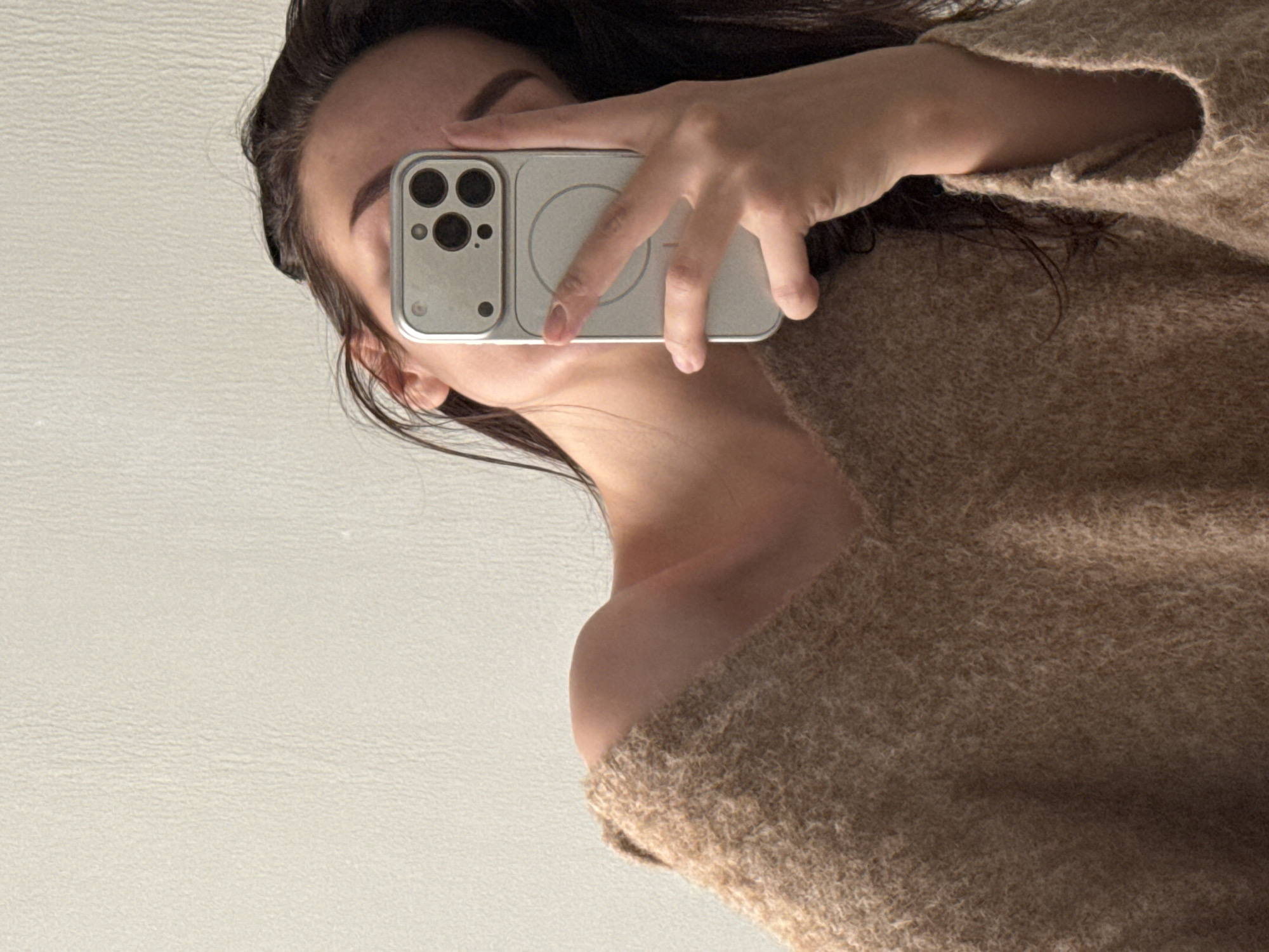 A person holds a silver iPhone 14 Pro with three camera lenses up to their face, partially obscuring it. They are wearing a beige, off-the-shoulder knit sweater. The background is a plain, light-colored wall. No information indicates this image was taken in Chaikovsky, Russia.