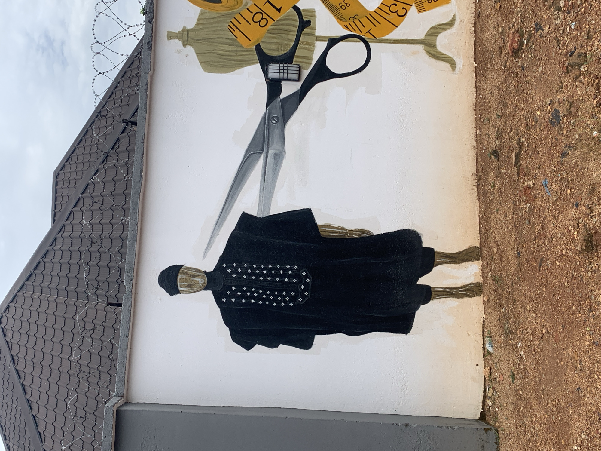 The image shows a mural on a white wall in Gura Top, Nigeria. The mural depicts a mannequin wearing traditional African attire, with a close-up of large scissors cutting through a golden measuring tape that is wrapped around a dress form. The mannequin is dressed in a black, long-sleeved garment with intricate white patterns on the chest and a black head covering. The scissors are large and metallic, with black handles, and appear to be cutting the measuring tape just above a segment marked with the number "18". The dress form is a muted gold color. Behind the mural, a building with a dark tiled roof and barbed wire along the top of the wall is visible. The ground in front of the wall is covered in red-brown gravel and small stones, with some scattered debris. The sky is overcast.