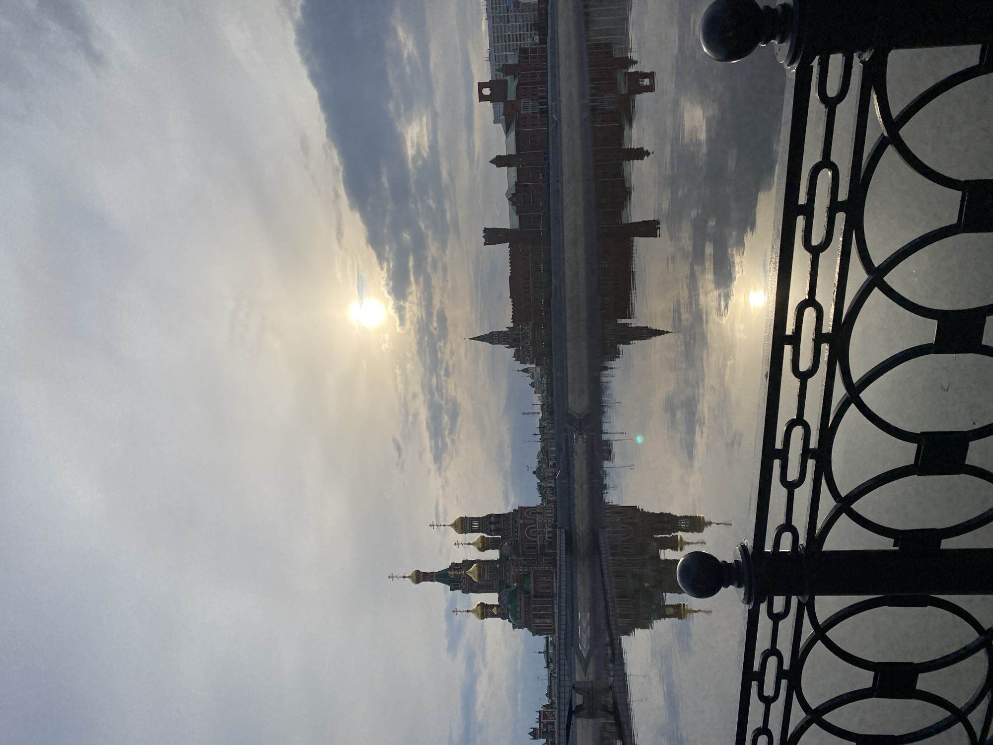 The image, when viewed upright, displays a serene cityscape in Yoshkar-Ola, Russia, characterized by prominent architectural structures reflected in calm water under a partly cloudy sky. In the foreground on the right, a decorative black metal railing with intricate circular and chain-link patterns partially frames the scene, suggesting the vantage point is from a bridge or waterfront promenade.
The left and central parts of the image are dominated by a collection of distinctive buildings. To the left, a Russian Orthodox-style church stands out with its multiple golden onion domes, topped with crosses, and dark red facades. Adjacent to it and stretching towards the right, a series of brick buildings with varying towers and turret-like features, reminiscent of medieval or European-inspired architecture, line the waterfront. These structures, along with the church, are almost perfectly mirrored in the tranquil water, creating a striking symmetrical composition.
The sun is visible low in the sky towards the left, partially obscured by soft clouds, indicating either sunrise or sunset. Its bright presence casts a warm glow across the scene and creates a strong, luminous reflection on the water. The sky above is a blend of light grey and blue with scattered clouds. No discernible human activity or visible text is present in the image, allowing the focus to remain on the architectural details and the beautiful interplay of light and reflections.