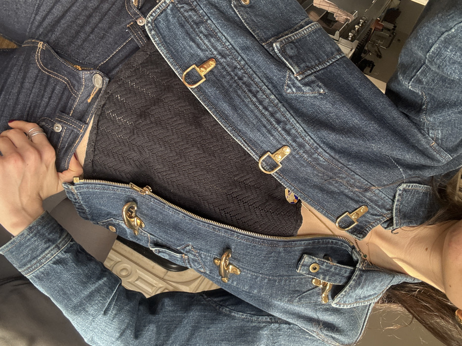 The image presents a close-up, eye-level view of a person's upper torso and arm in an indoor setting. The subject is wearing a dark blue denim jacket featuring five prominent gold-colored toggle-style fasteners and chest flap pockets, unzipped to reveal a black, patterned or knitted camisole underneath. Dark blue denim jeans, partially unbuttoned and unzipped, are visible at the bottom of the frame. The person's bare right hand, adorned with two silver-toned rings on one finger and dark-painted fingernails, rests casually near the waistband of the jeans. A thin necklace with a small pendant is visible on the neck, and a portion of a gold earring is seen near the right collarbone. The background is largely out of focus, but hints of a blurred cityscape are visible through a window in the upper left, while a distinct light-colored, ornate architectural detail, resembling a fireplace mantelpiece or decorative wall molding, occupies the mid-right. The ambient lighting appears natural and bright, indicating a daytime scene. The architectural and background elements are consistent with a residential interior in New York City, United States of America.