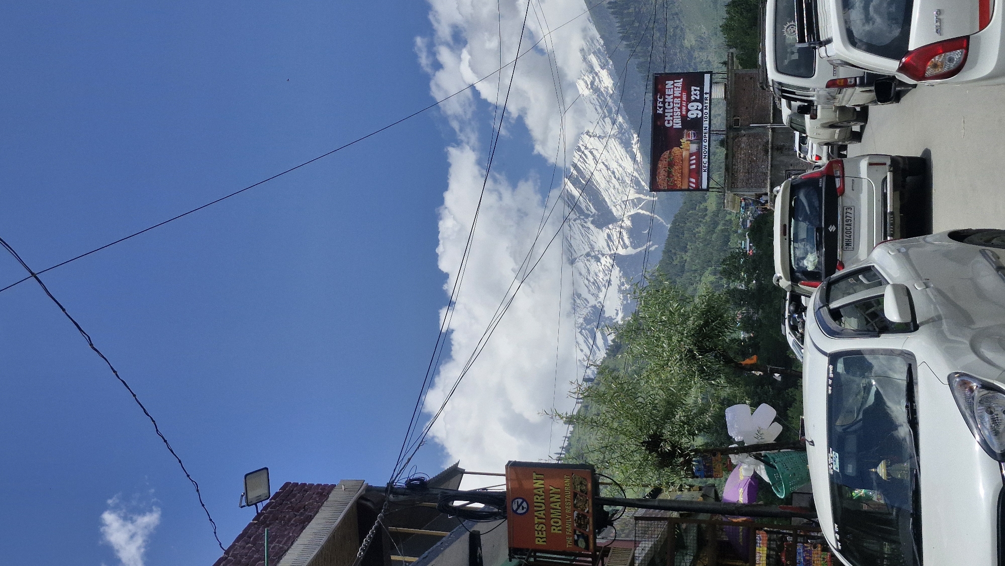 The image shows a street scene in Bahang, India, with a clear blue sky and snow-capped mountains in the background. A variety of vehicles are present on the road, including white cars and SUVs. A prominent sign for "RESTAURANT ROMANY" is visible on the left side of the image, along with another sign advertising a "KFC CHICKEN KRISPER MEAL." Power lines crisscross the sky, and trees and buildings line the street. The overall atmosphere suggests a busy, sunny day in a mountainous region.