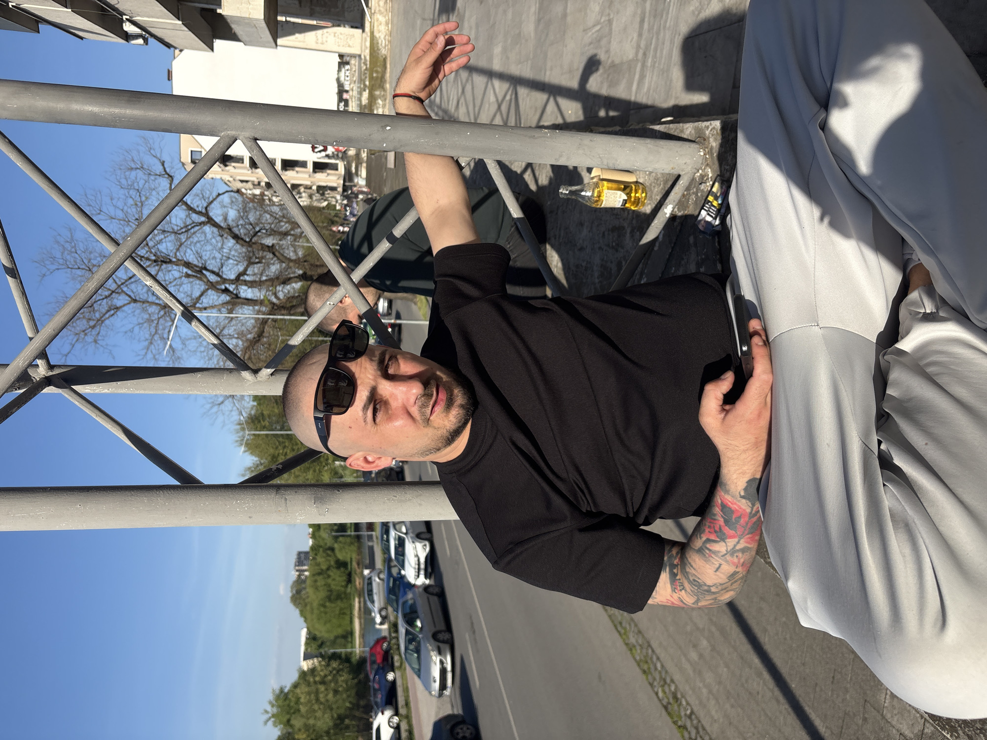 The media file presents an outdoor scene in an urban environment, captured from a low angle and rotated 90 degrees counter-clockwise. A prominent gray metal railing forms a significant part of the foreground and midground, suggesting the subjects are on an elevated structure like a balcony or overpass.
The primary subject is a man with a shaved head, stubble, and dark sunglasses, looking directly at the camera with a neutral expression. He wears a black short-sleeved t-shirt, showcasing a heavily tattooed left arm with red and black designs. His right arm is bent and rests on the railing.
To the right, partially visible, are the light gray pants and legs of what appears to be a second person, holding a smartphone. Between the two, a small clear glass bottle containing yellow liquid (likely alcohol) and a small, colorful packet (possibly cigarettes) are placed on the ground or a ledge.
The background reveals a clear blue sky, indicating bright daylight, likely midday or afternoon, given the strong shadows cast. Beyond the railing, a street with parked cars is visible. Various buildings, some seemingly residential or commercial, and trees (both bare and with green foliage) extend into the distance. A partial sign on one building in the background reads "BECK'S" and "BEERS."
The image captures a casual, relaxed moment in an urban setting, consistent with Plovdiv, Bulgaria.