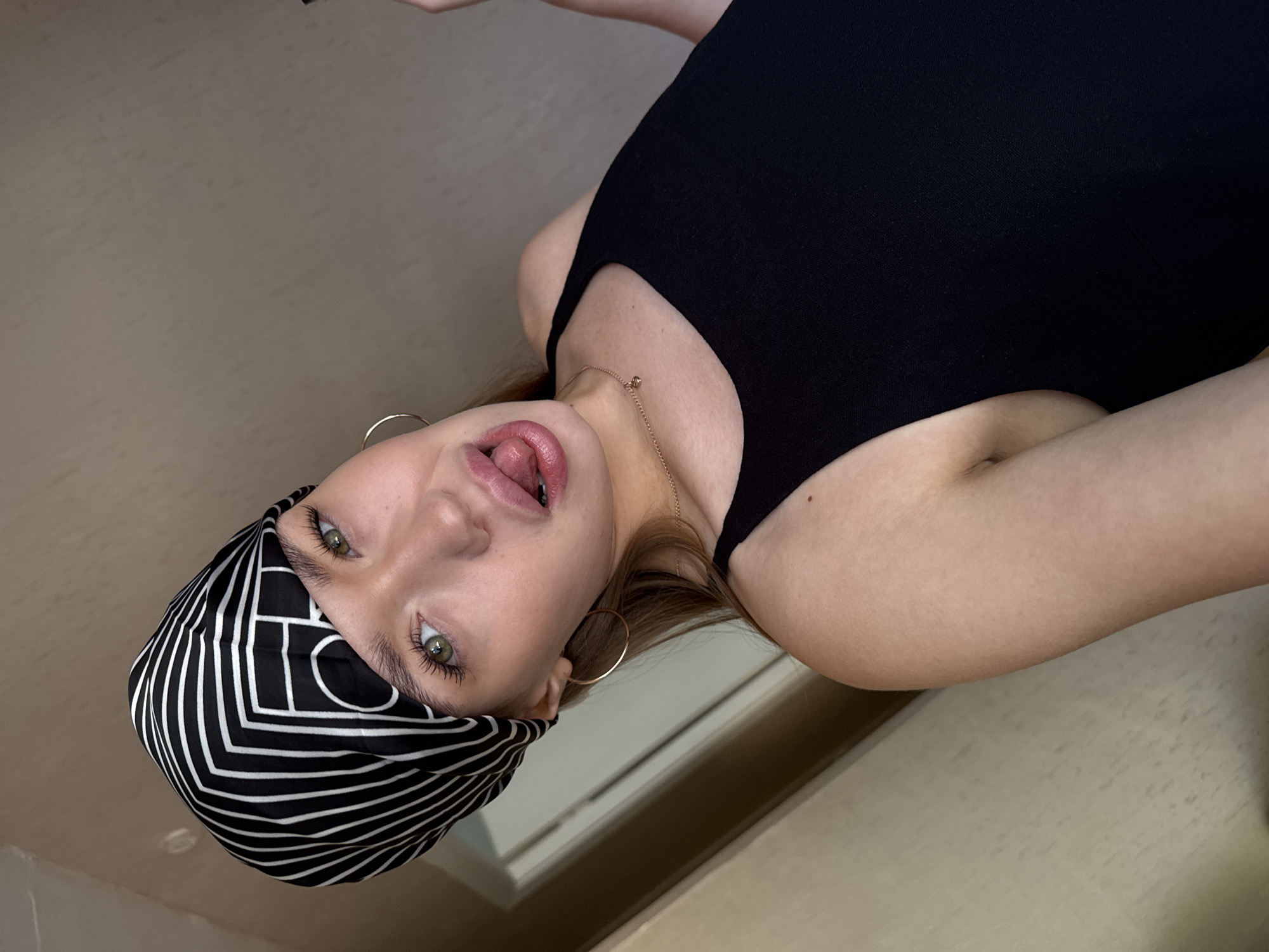 A young woman with green eyes and blonde hair is looking at the camera and sticking her tongue out. She is wearing a black tank top and a black and white patterned headscarf. She has gold hoop earrings and a gold necklace with a small heart pendant. The background is a plain wall. The image is taken indoors. It is not possible to determine the location in the city of Chaikovsky, Russia from the provided image.