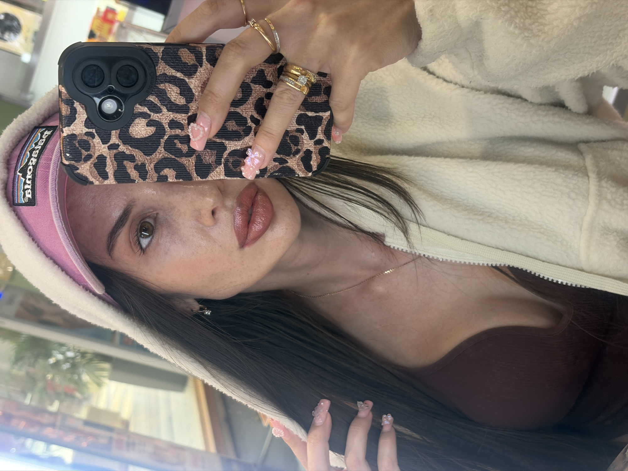 The image shows a woman taking a selfie. She is wearing a pink baseball cap with a Patagonia logo and a white fluffy zip-up jacket. She has long, dark hair and is wearing makeup, including lipstick and nail polish. Her fingernails are decorated with small flowers and pearls. She is also wearing several gold rings on her fingers, including one with diamonds. The phone she is holding has a leopard print case. The background appears to be indoors, with some blurred reflections visible. The lighting suggests it is daytime. The woman's expression is neutral, and she seems to be posing for the photo.