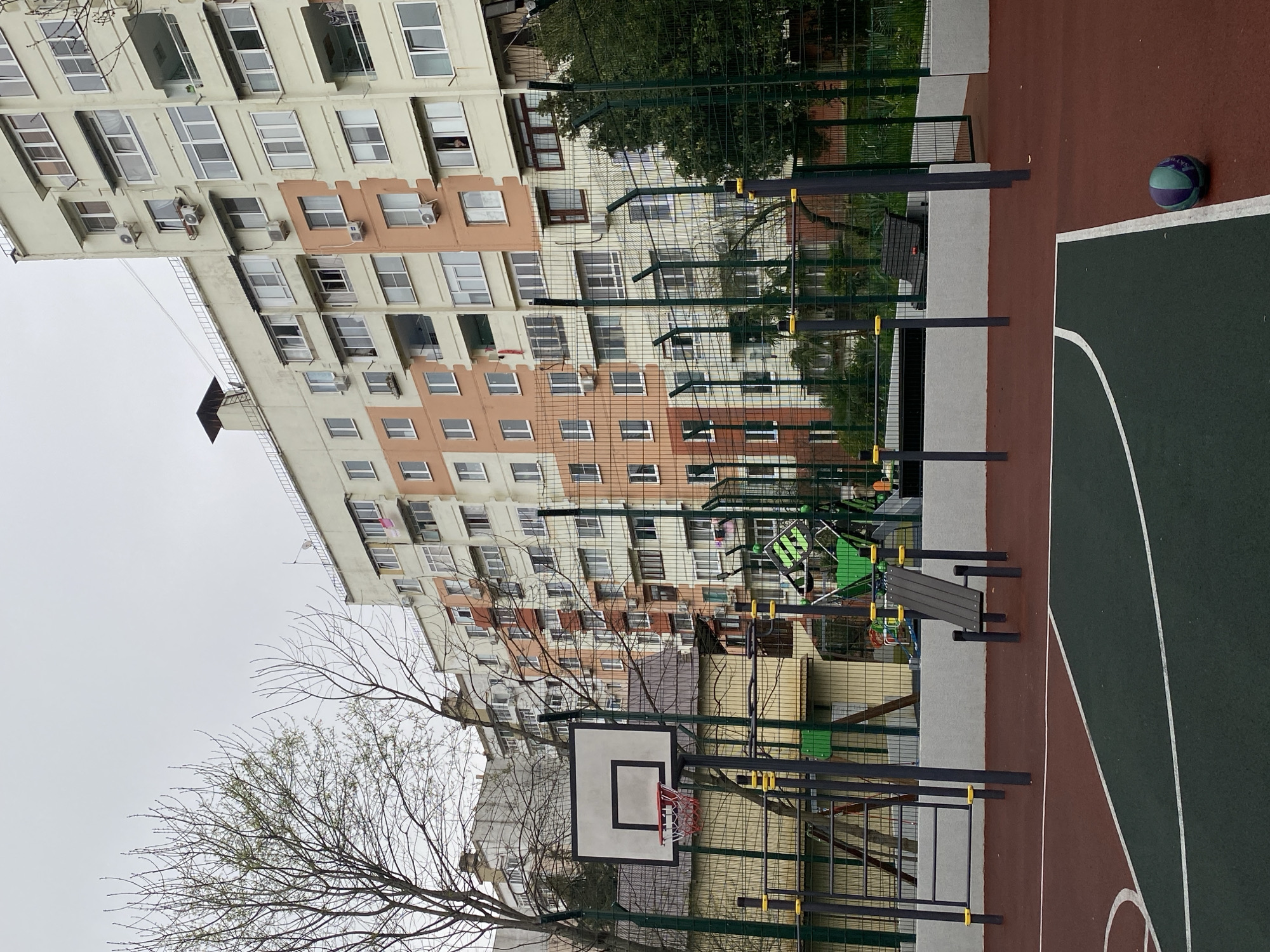 The image shows an outdoor basketball court next to an apartment building. The court is green and maroon with white lines. A basketball rests on the court's edge. Behind the court is a playground with climbing equipment and a bench, surrounded by a green fence. A basketball hoop stands to the left. The apartment building in the background is a high-rise with cream and peach colored sections. It is a cloudy day. While not visible in the image, this scene is reminiscent of the city of Sochi, Russia.