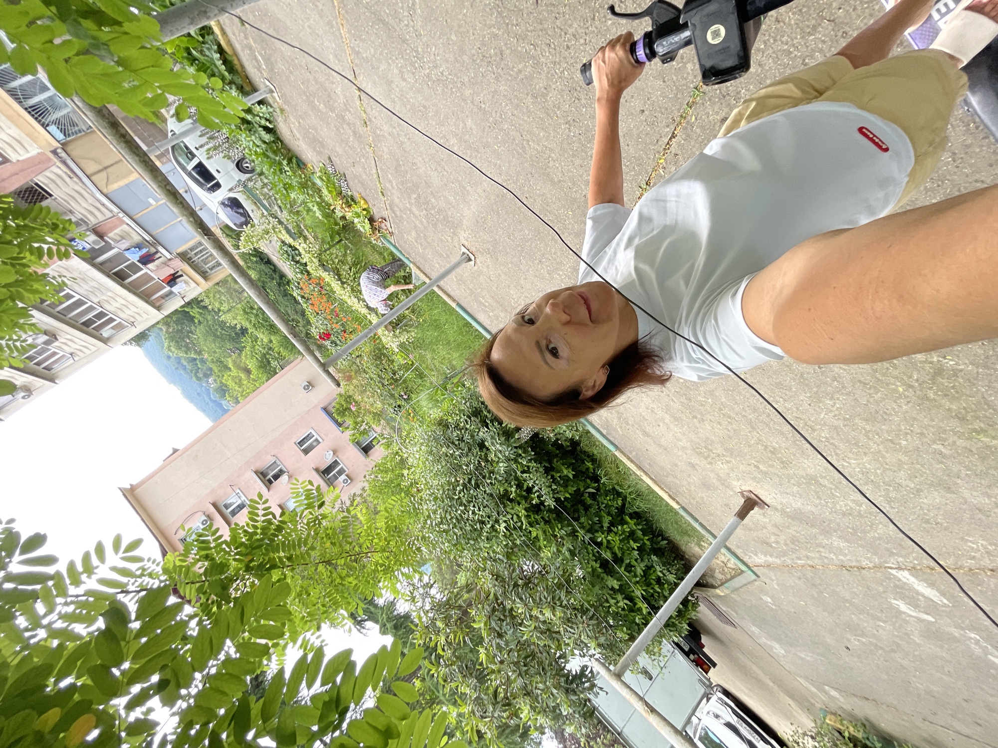 The image depicts an outdoor scene with a woman holding the handlebars of a scooter or electric bike. She is wearing a white t-shirt and khaki shorts. In the background, there are apartment buildings, trees, and a paved area. A white car is parked in the background. The scene appears to be in an urban residential area, possibly in Sochi, Russia, given the architecture of the buildings and the vegetation. The orientation of the image is rotated, making it appear as if the ground is vertical.