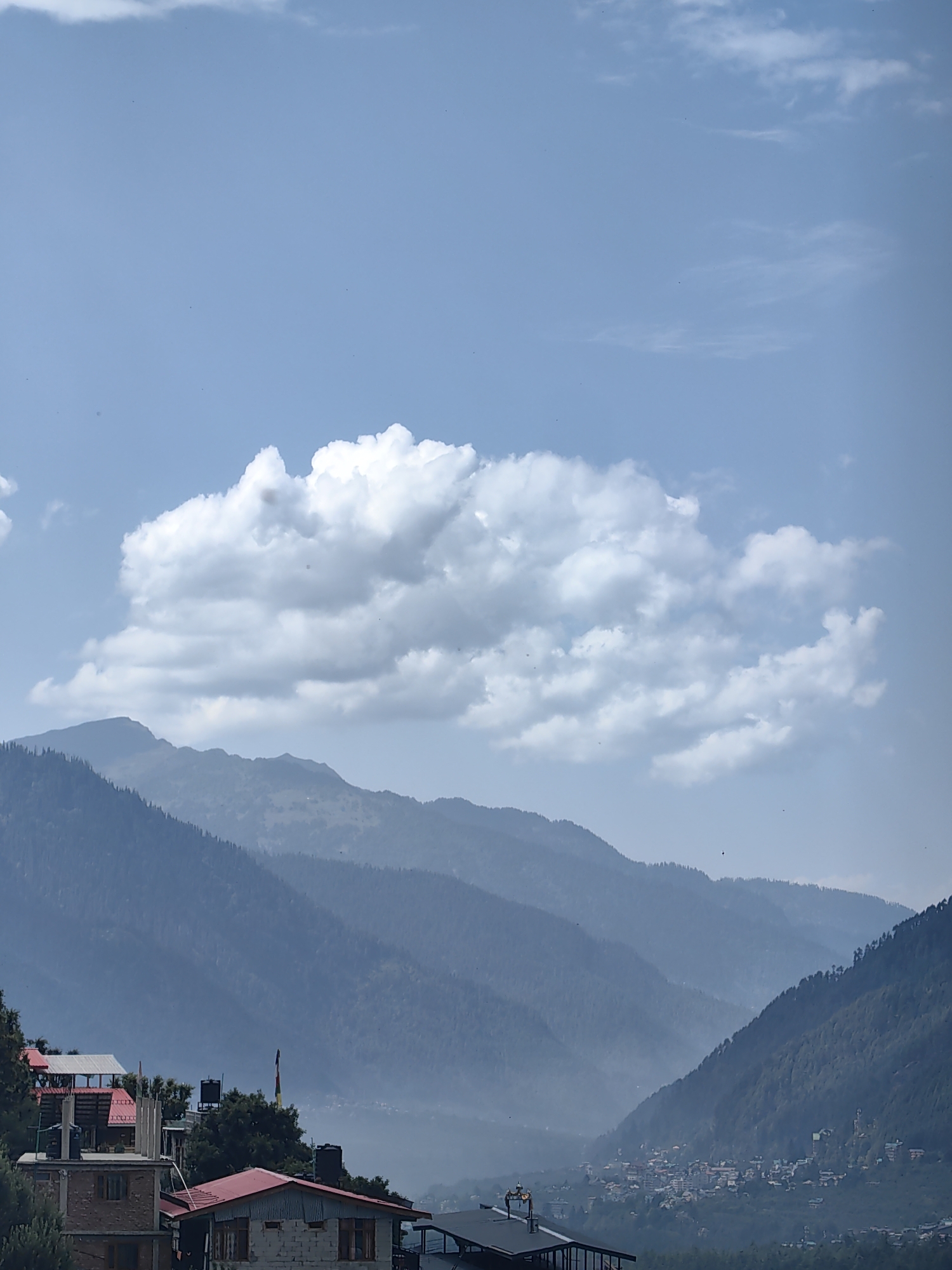 The image depicts a mountainous landscape in Vashisht, India, under a partly cloudy sky. In the foreground, several buildings with red tiled roofs are visible, some appearing to be multi-story residences or guesthouses. These structures are nestled amongst lush green trees.
The mid-ground is dominated by a valley with a small town or village situated at its base. The town is densely packed with buildings, suggesting a populated area. The valley is shrouded in a light atmospheric haze, which softens the distant view.
The background features layers of steep, tree-covered mountains that recede into the distance. The mountains exhibit varying shades of blue and green, indicating depth and the effect of atmospheric perspective. The sky is a pale blue with scattered white cumulus clouds, one large cloud formation being particularly prominent in the upper half of the image. There are no visible people or active events captured in the scene. The overall impression is one of a serene, natural environment with human settlements integrated into the mountainous terrain.