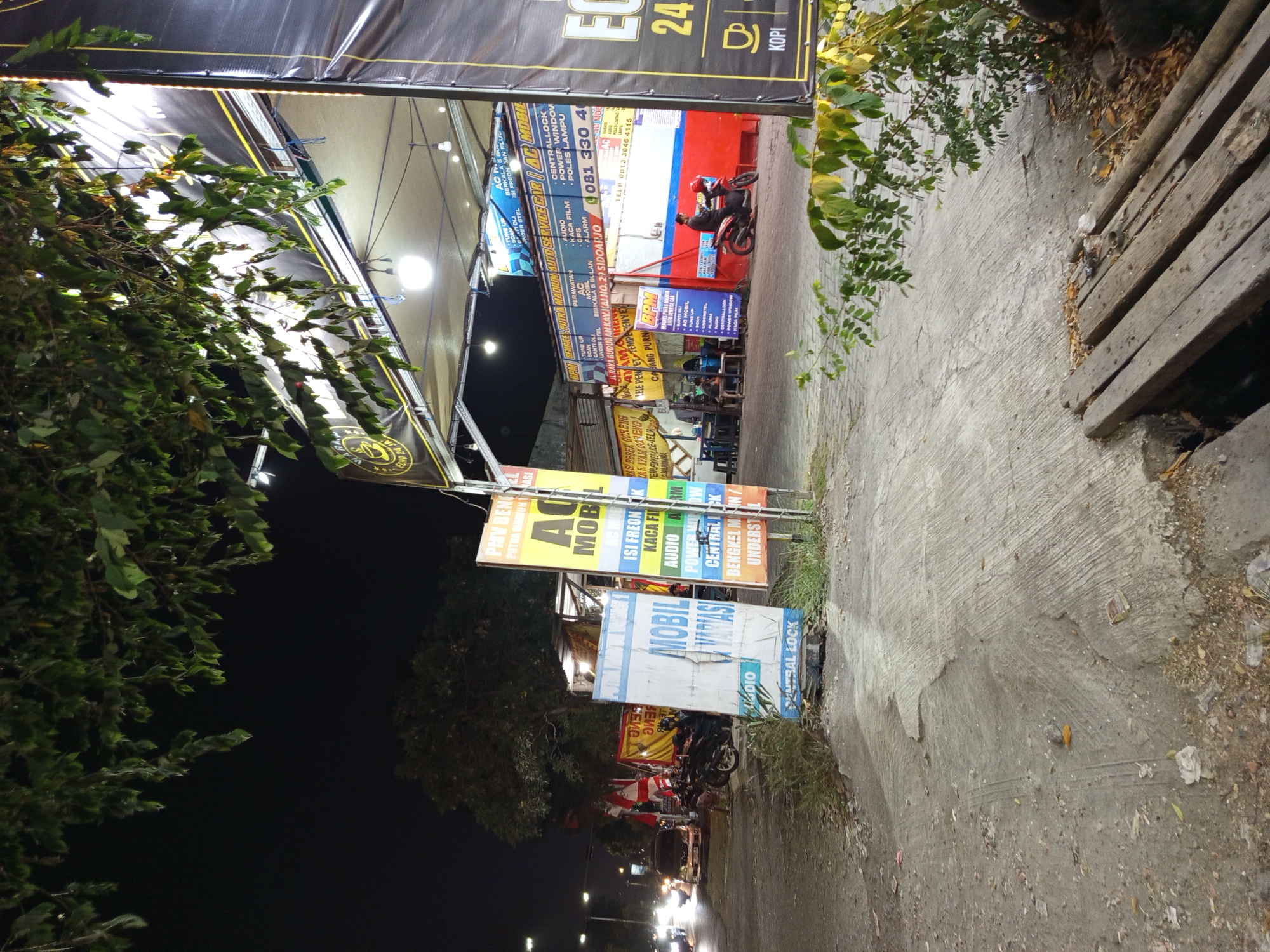 The image depicts a street scene at night in Buduran, Indonesia, near an auto service shop. A large, black banner with yellow lettering, partially obscured by trees and foliage, hangs on the right side of the frame, advertising "KOPI" (coffee) and "24" with a coffee cup icon. To the left, a multi-story building houses an auto service center, indicated by signs such as "BPM AUTO SERVICE CAR" and "AC MOBIL". Various services like "TUNE UP", "ISI FREON", "KACA FILM", "AUDIO", and "CENTRAL LOCK" are advertised on colorful banners and signs.
In the foreground, a damaged white and blue sign with "MOBIL VARIASI" and "AUDIO CENTRAL LOCK" written on it lies tilted on the ground. A dirt and concrete road runs through the scene, with a few motorcycles parked on the side. A person wearing a red helmet is seen riding a motorcycle in front of the auto service shop. Other people are seated at tables inside the shop's open area. Cars are visible on the street in the background, with headlights illuminating the darkness. The environment appears to be a commercial area with elements of both residential and automotive services.