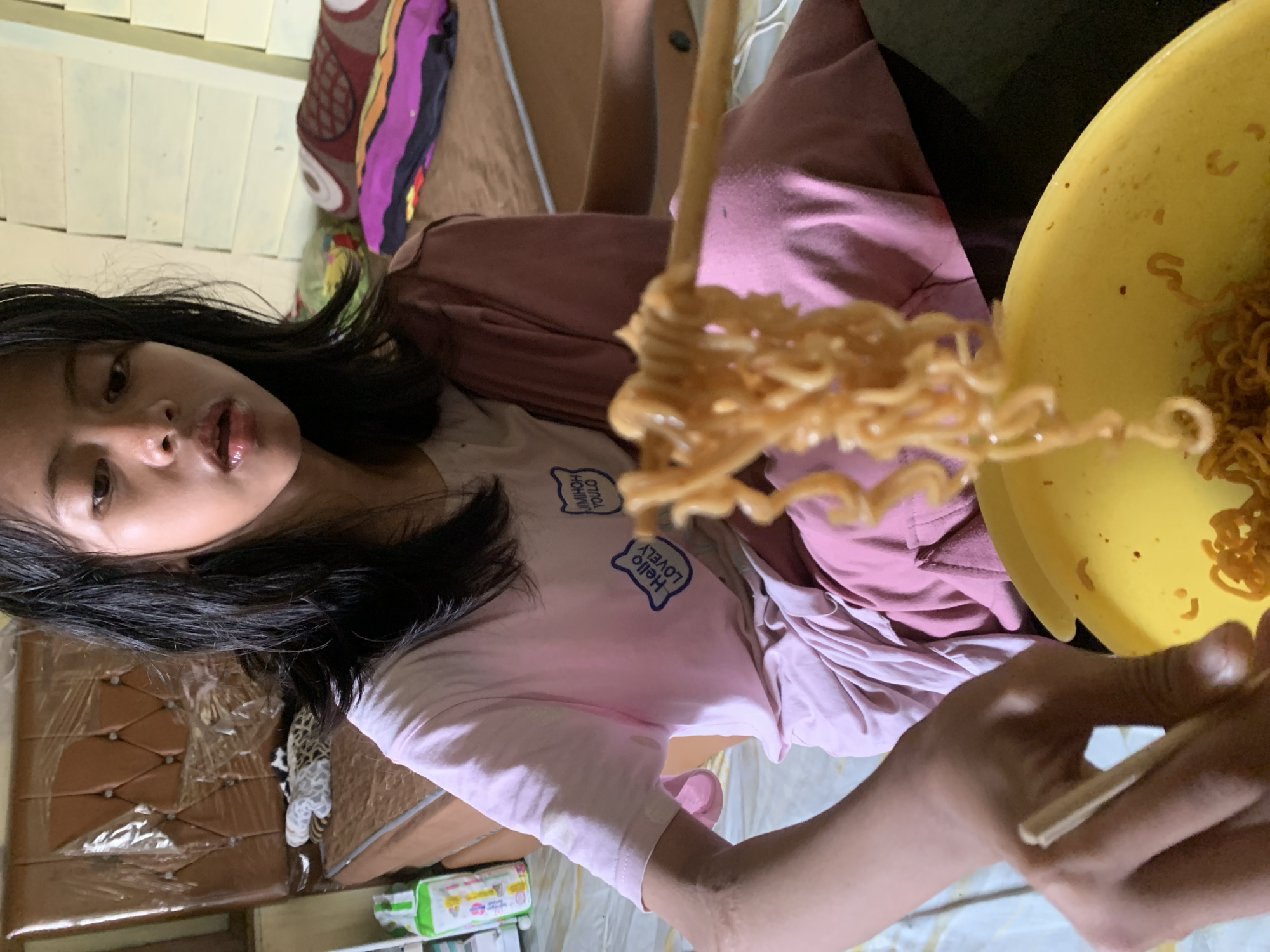 A young female with dark hair, wearing a pink t-shirt with graphics, is sitting on a bed and eating instant noodles with chopsticks. She is in a bedroom in Sungai Meriam, Indonesia. The bed has a brown headboard covered in plastic wrap, and there are pillows and a blanket on the bed. A yellow bowl of noodles is visible in the foreground.