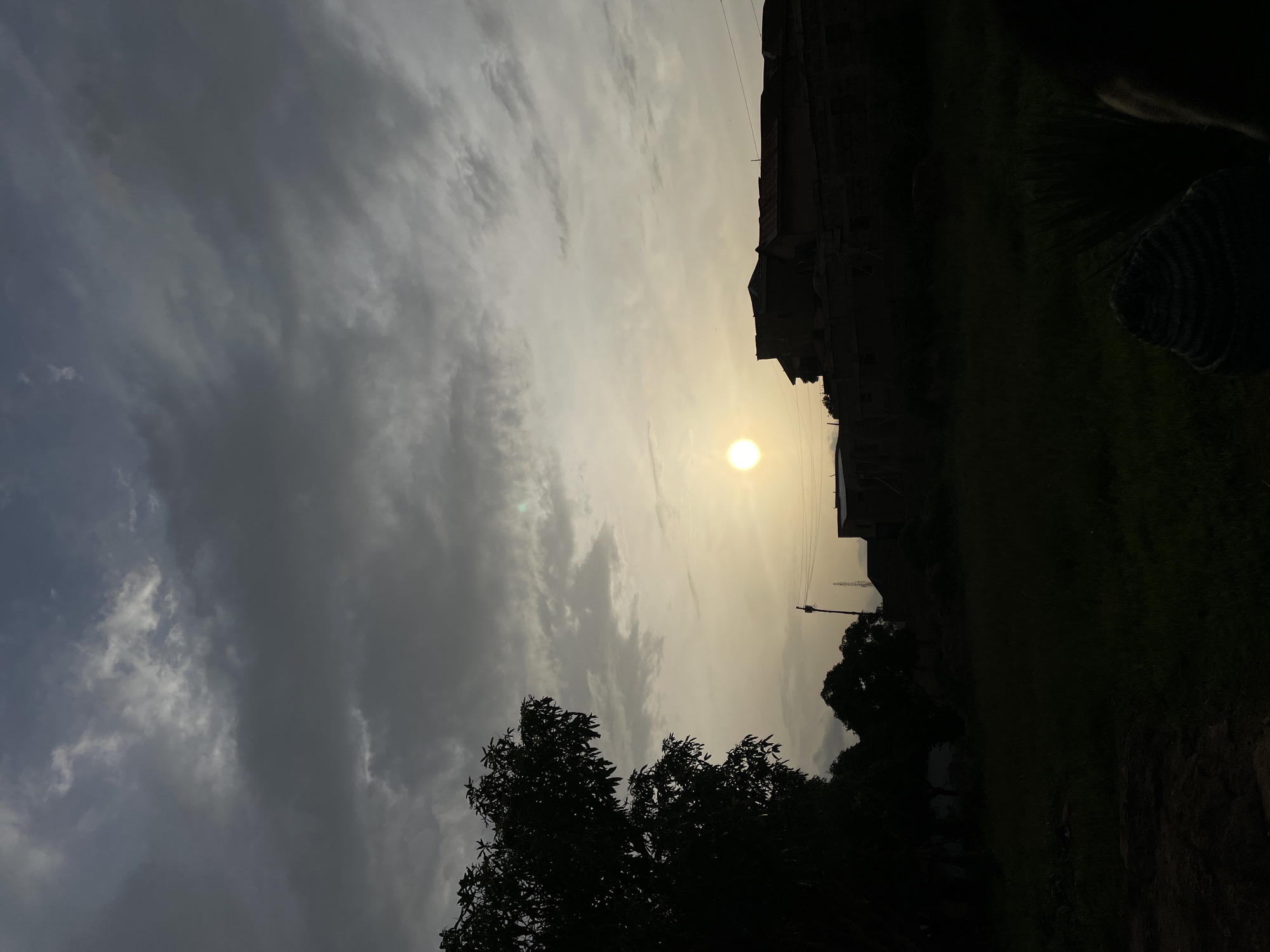 The image shows a cityscape during sunset in Jos, Nigeria. The sun is low in the sky, partially obscured by clouds, casting a warm glow over the scene. In the foreground, silhouetted trees stand against the sky. Buildings and structures are visible in the middle ground, appearing as dark outlines against the lighter sky. Power lines are also visible. The overall impression is of a city landscape bathed in the light of the setting sun, with a mix of natural and built environments.