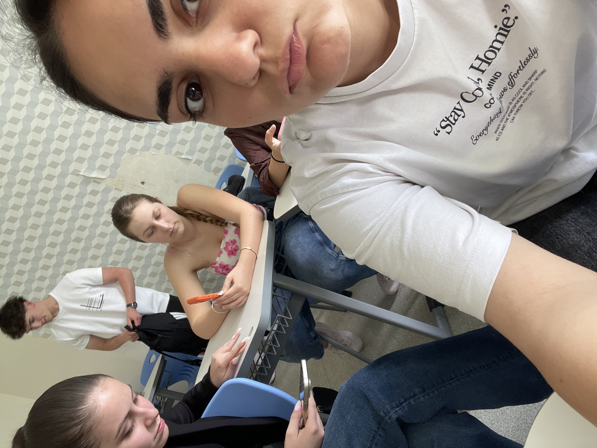This selfie-style photo, captured in what appears to be a classroom or study area in Burgas, Bulgaria, features several young individuals. The primary subject is a young woman taking the photo, her face and upper body dominating the foreground. She has dark hair, expressive eyebrows, and a somewhat serious expression, looking directly into the camera. She wears a white long-sleeved t-shirt with black text, part of which reads, "Stay Cool, Homie." and includes phrases like "Everything flows effortlessly" and a motivational quote about having a cool and sharp mind.
In the background, other young people are visible. To the right, another young woman with braided hair wears a floral top and is seated at a desk, looking down. In the mid-ground, a young man in a white t-shirt stands or leans, holding a black backpack. Further to the left foreground, another young woman with dark hair, dressed in a dark top, is also seated at a desk, seemingly engrossed in her phone.
The setting is an indoor space with light-colored walls adorned with a repeating geometric cube pattern. There are some signs of wear on the wallpaper. Standard classroom-style desks and chairs with blue seats are present. The overall atmosphere appears casual and relaxed, with individuals engaged in their personal activities. The lighting suggests it is daytime. Objects like phones (one orange, one silver/white) are visible in the hands of the individuals, hinting at a modern, connected environment.