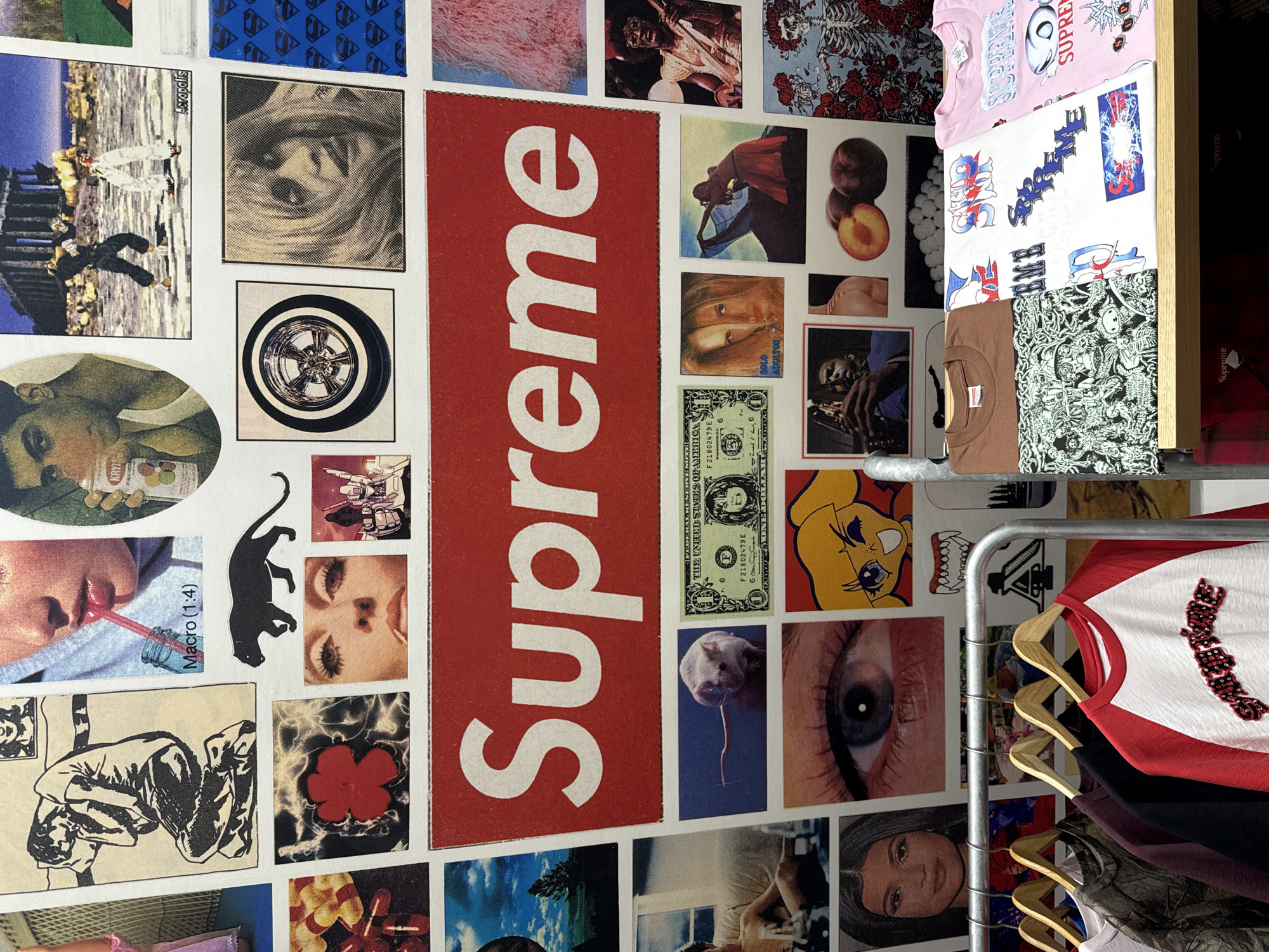 This image depicts an interior scene within a Supreme retail store, located in New York City, United States of America. The primary visual element is a wall completely covered with a dense, eclectic collage of various printed images. Central to this collage is a large, rectangular red sticker or poster bearing the word "Supreme" in white, bold, uppercase letters.
The surrounding collage includes a diverse array of subjects:
- **People:** Portraits of individuals, including Kylie Jenner, a man holding a drink, a woman drinking through a straw, a woman with long hair (labeled "SOLO ADULTOS"), and a musician playing a saxophone.
- **Art/Illustrations:** A black and white sketch of a crouching figure, a black silhouette of a panther, an anime-style character, a robot, and a detailed illustration of skulls and roses.
- **Objects:** A US dollar bill, a stylized wheel rim, two peaches, and a red flower with a luminous border effect.
- **Animals:** A small white mouse.
- **Scenes:** Two figures engaged in combat in front of what appears to be the Parthenon ("Acropolis" is printed below this image).
- **Abstract/Textures:** A close-up of an eye, a pink fluffy texture, and a pattern of blue squares containing Superman 'S' shield logos.
Below the wall collage, a retail display area is visible. On the left, a silver-colored metal clothing rack holds multiple garments on wooden hangers, including a white baseball-style shirt with red raglan sleeves and a dark graphic design on the chest, a dark purple long-sleeved shirt, a black long-sleeved shirt, and a camouflage-patterned shirt. On the right, a light-colored wooden shelf displays several folded t-shirts, including a plain brown shirt, a white shirt with an intricate black and white graphic pattern featuring skulls and figures, another white shirt adorned with various colorful "Supreme" logos and graphics, and a pink shirt also bearing "Supreme" branding and additional designs. A light-colored floor, possibly concrete, is visible beneath the displays.