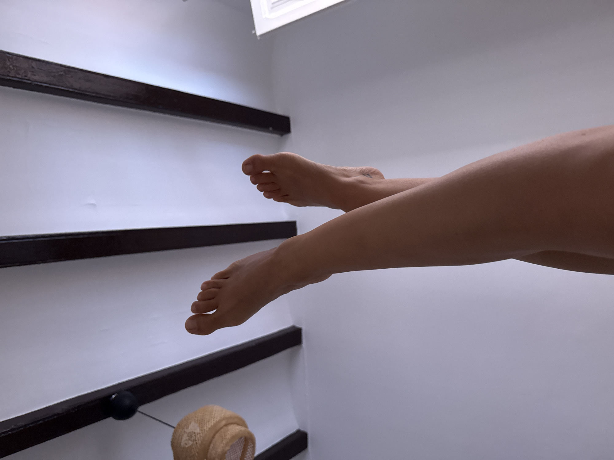The image shows a pair of bare feet and legs of a person suspended against a white ceiling with dark wooden beams. A woven light fixture hangs from the ceiling on the left side. The perspective is from below, looking up. The scene does not provide enough information to determine a specific location in Barcelona, Spain.