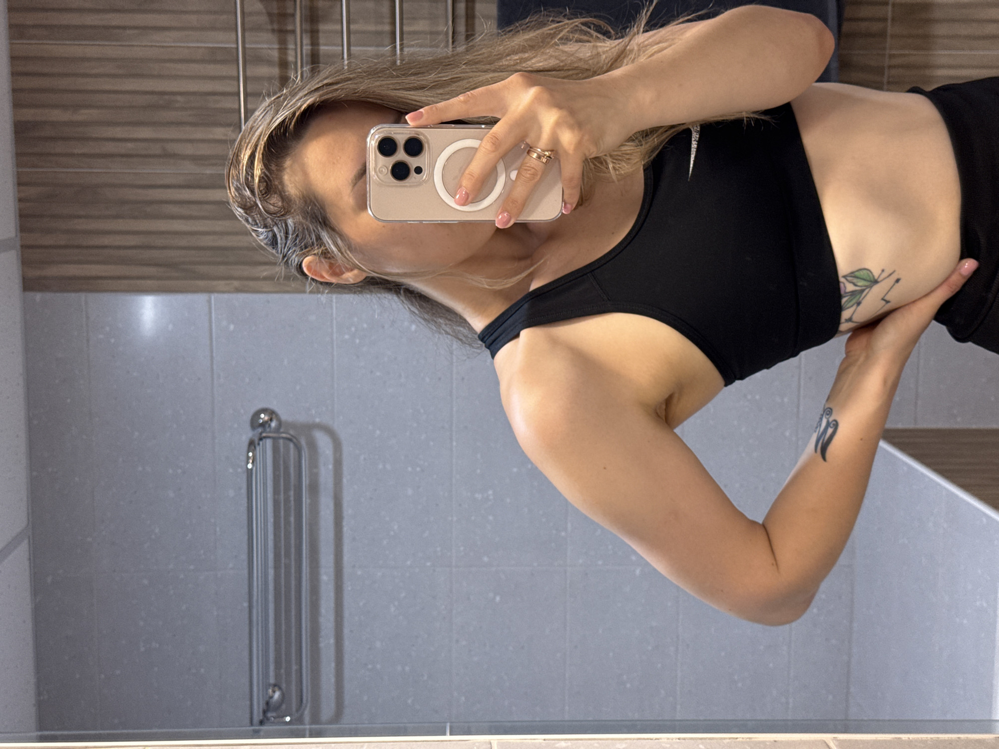 A woman in a black sports bra and black athletic shorts is taking a mirror selfie in a bathroom. She has long blonde hair and is wearing rings on her left hand. There are tattoos on her left arm and ribs. The bathroom has gray tiled walls and a wooden accent wall. A towel rack is visible on the wall.