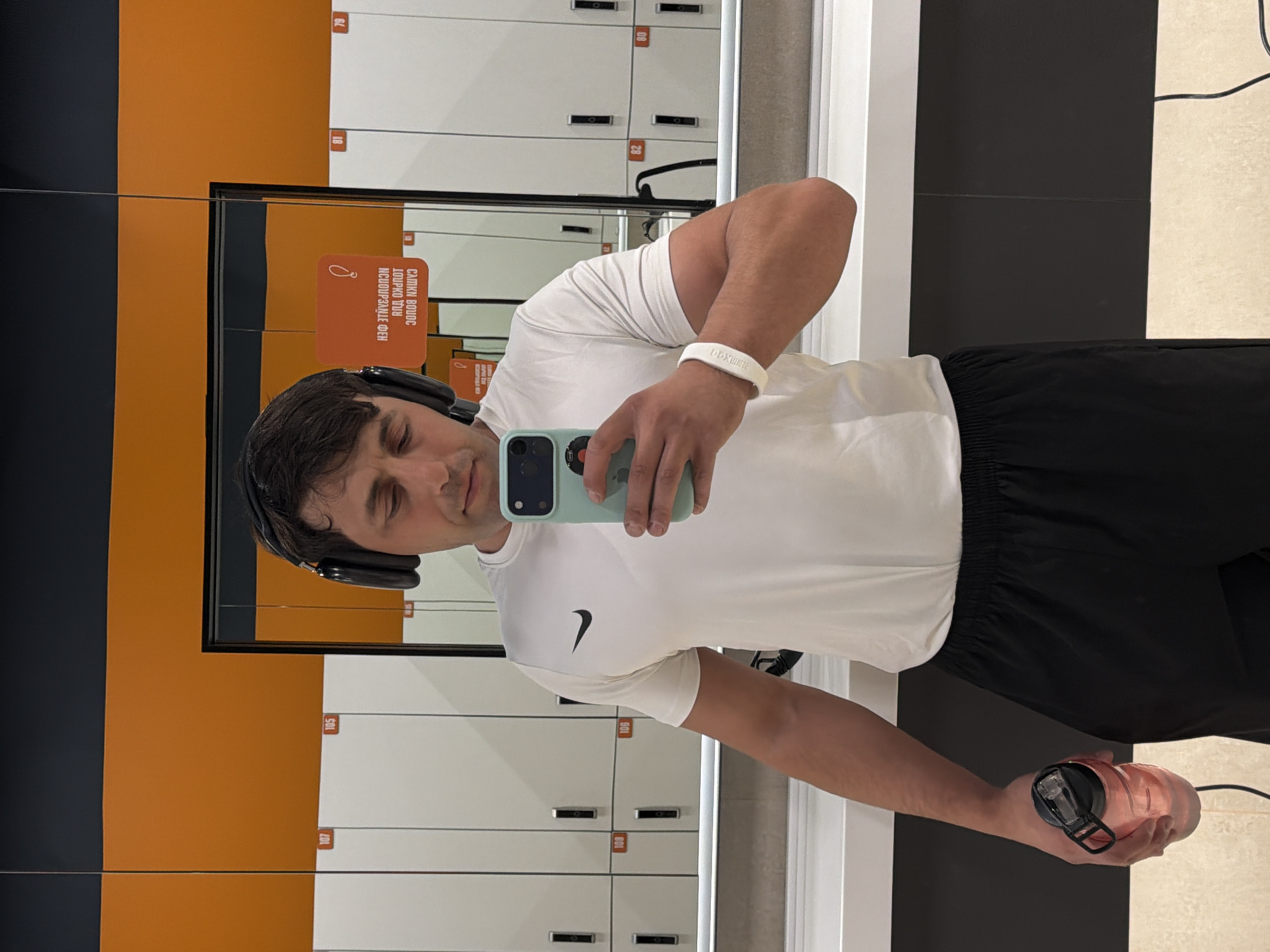 A man is captured in a mirror selfie, likely within a locker room or changing area of a gym. He is seen from the chest up, with his eyes partially closed in a relaxed or slightly self-conscious expression. He wears a white short-sleeved t-shirt with a black Nike swoosh logo on the left chest, black shorts, and black over-ear headphones. A white wristband, possibly bearing the logo "DDXWorld" or a similar gym name, is visible on his right wrist.
In his right hand, he holds a light blue smartphone with a green case, which is reflecting his face as he takes the picture. His left hand holds a translucent rose gold or pink water bottle with a black lid and carrying loop.
The background, largely reflected in the mirror, consists of a row of white lockers with dark-colored handles or locks. Several locker numbers are visible in red, such as 79, 81, 105, 106, 107, and 108, appearing reversed due to the reflection. Behind the lockers, a wall is visible with distinct orange and black sections. An orange rectangular sign is reflected in the mirror, displaying Cyrillic text which includes the phrase "ИСПОЛЬЗОВАНИЕ ФЕН" (use hairdryer) and other less decipherable words that appear to relate to gym rules or amenities.
The scene suggests an indoor fitness environment, possibly in Krasnoyarsk, Russia, given the Cyrillic text and potential gym chain identification. The man appears to be pausing for a photo either before or after a workout.