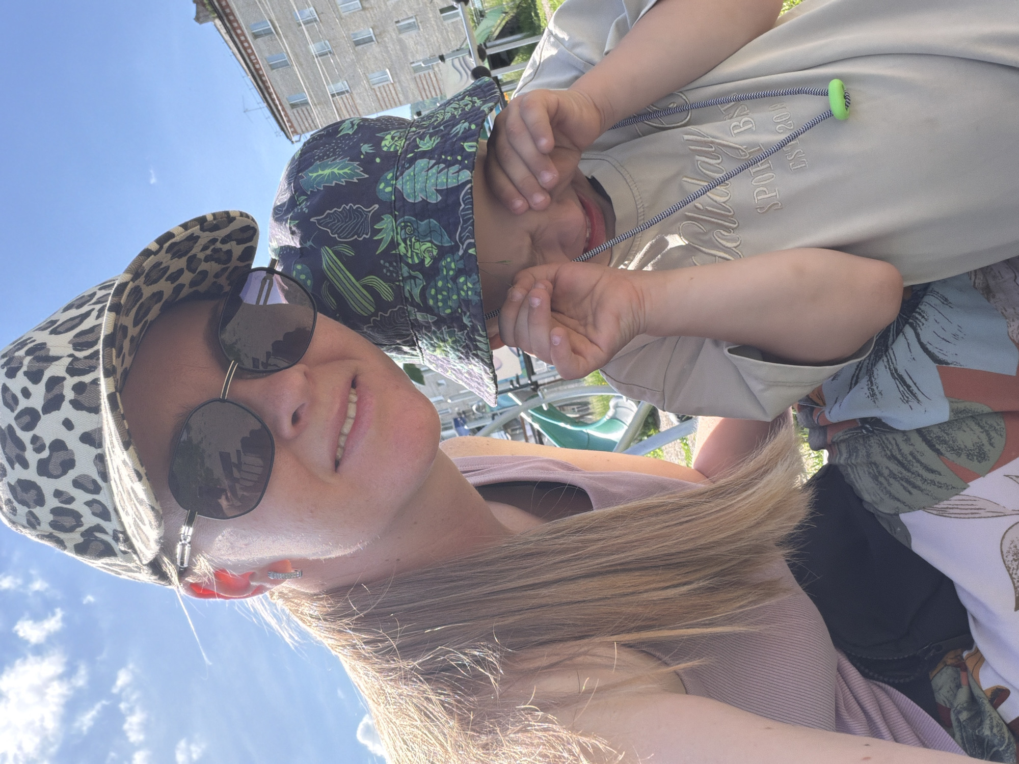 A woman and a child are taking a selfie outdoors on a sunny day, with a residential building and playground equipment visible in the background. The woman, on the left, is wearing a leopard-print baseball cap, sunglasses, and a pink tank top. Her blonde hair is long and loose. The child, on the right, is wearing a bucket hat with a cactus print and a light-colored t-shirt. The child is rubbing their eyes. They appear to be sitting on a blanket with a floral pattern. The location is identified as being in the city of Kuldur, Russia.