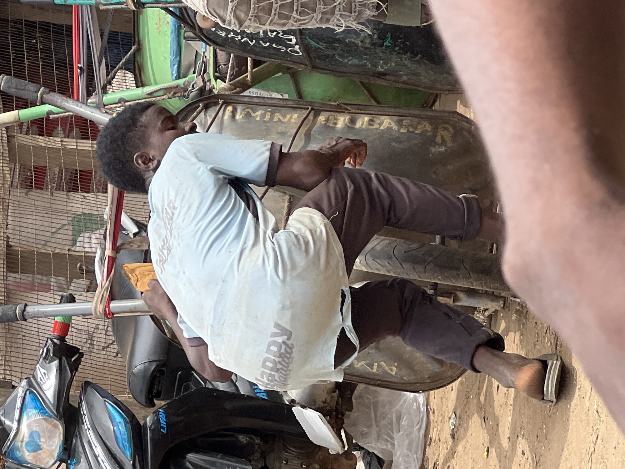 In Oko Amakom, Nigeria, a young Black male, wearing a torn light blue t-shirt with the faded imprint of "Happy Matrid" and dark brown pants, is seen from behind. He is hunched over a motorcycle, with his left hand resting on the handlebars and his right hand near a piece of bread he holds. The motorcycle is black and partially visible. To the right of this motorcycle, another motorcycle with a green body and faded yellow lettering is present, along with a third motorcycle, partially obscured, that has torn white fabric draped over its side. The ground is sandy and brown, with some debris scattered around. The scene appears to be outdoors, possibly in a market or roadside setting, indicated by the presence of multiple motorcycles and what looks like a fence in the background.