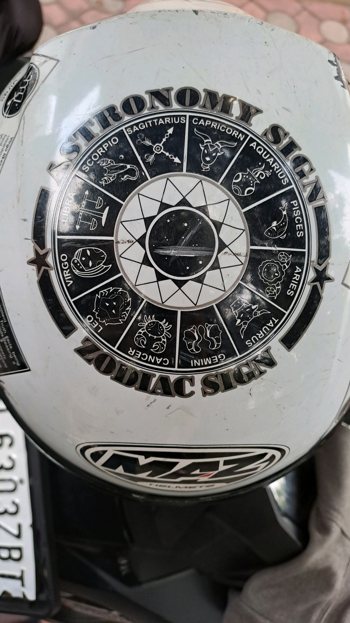 The image shows a close-up of a white helmet with black and silver graphics. The graphics depict a circular zodiac wheel with symbols for Sagittarius, Capricorn, Aquarius, Pisces, Aries, Taurus, Gemini, Cancer, Leo, Virgo, Libra, and Scorpio. The words "ASTRONOMY SIGNS" and "ZODIAC SIGNS" are also visible. Below the zodiac wheel, the logo "MAZ HELMETS" is present. The helmet is resting on a dark surface, possibly a motorcycle seat, and a portion of a license plate with the partial inscription "6303ZBT" is visible on the left side. The background is blurred but appears to be outdoors with a reddish-brown textured surface, suggesting a paved area. The overall condition of the helmet shows signs of wear and tear, with scuffs and scratches on its surface. The image lacks any discernible geographical landmarks that would specifically place it in Julicotmeurak, Indonesia, or any other specific city. There are no people or actions depicted in the scene.