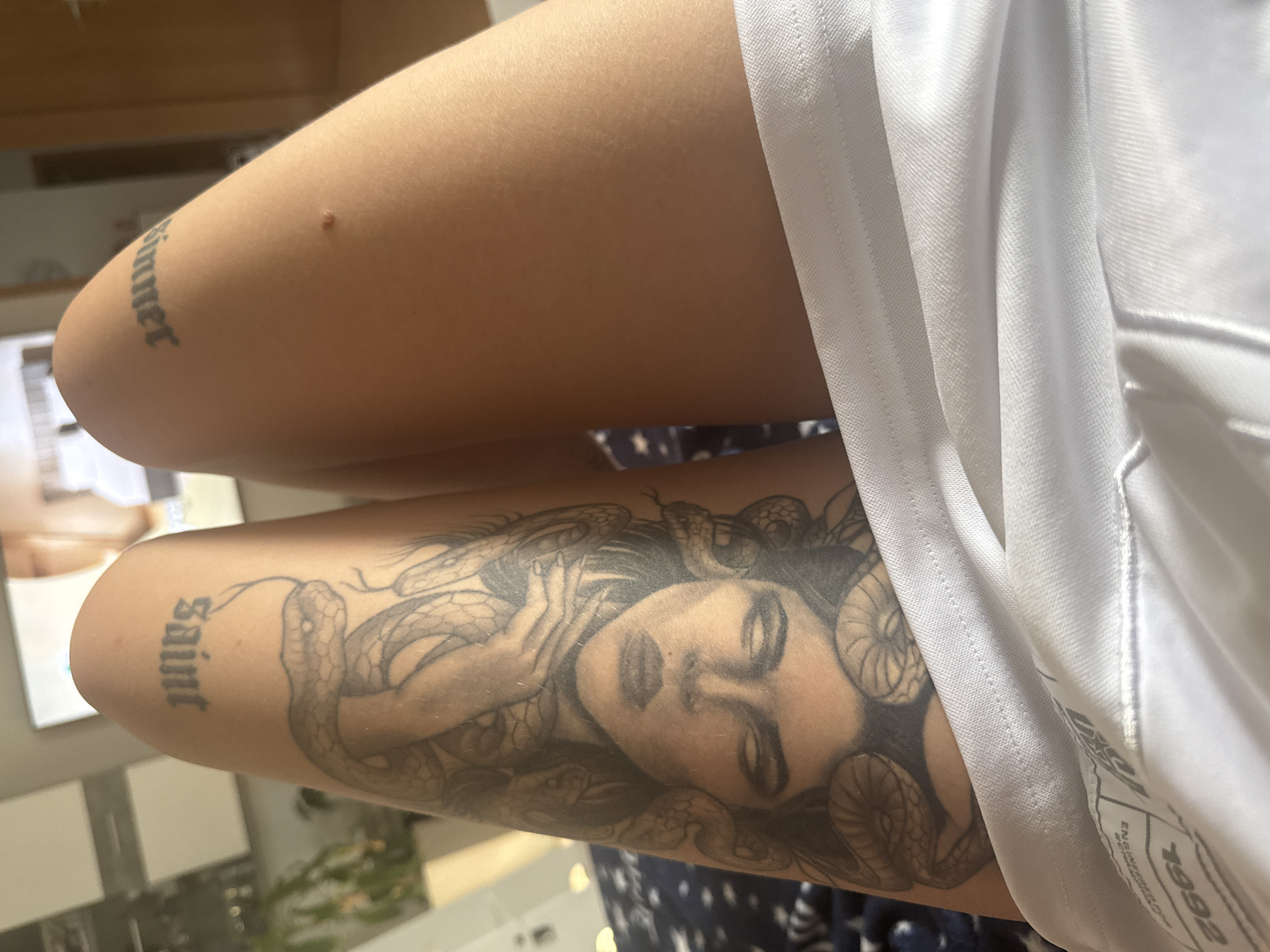 The image shows a close-up view of a person's thighs. The left thigh features a large tattoo of Medusa with snakes coiled around her head, and the word "Saint" in gothic font above it. The right thigh has the word "Sinner" in gothic font. The person is wearing white fabric, possibly shorts or underwear, that covers the lower part of their thighs. The background is blurred but appears to be an indoor setting with a television screen visible on the wall. There is a plant to the left and a doorway to the right. The lighting suggests it is daytime.