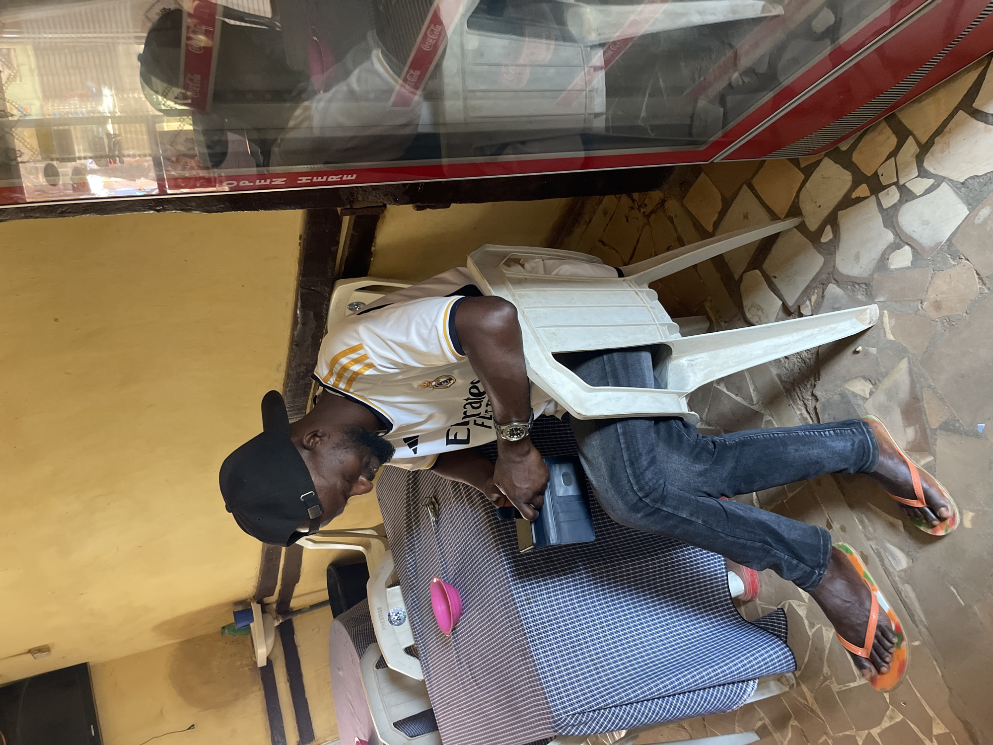 A man wearing a black cap, a white jersey with gold stripes and "Emirates FLY Better" lettering, and jeans, is sitting on a white plastic chair in Samaru, Nigeria. He is holding a dark gray device. His feet, adorned with flip-flops, are visible extending from the chair. The area features a tile-covered floor and yellow walls. A Coca-Cola cooler is visible overhead. Another chair and a blue and white checkered patterned mattress are also present.