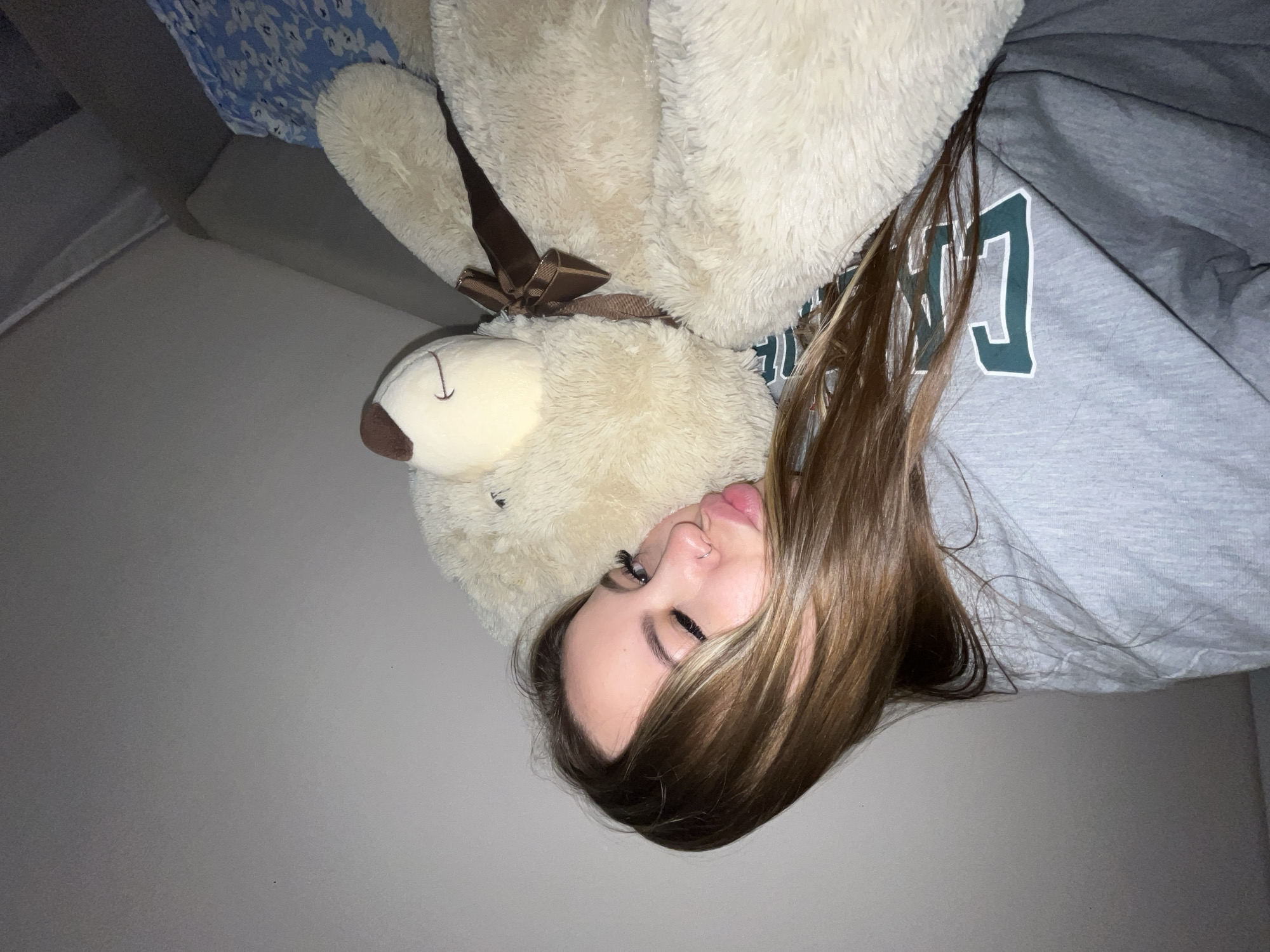 A young female, likely an adolescent or young adult, is depicted lying down, resting her head on or next to a large, cream-colored teddy bear. The image is oriented with the female's head to the lower right, implying a 90-degree counter-clockwise rotation from a natural upright position.
The female has long, light brown hair with subtle highlights, flowing around her head. Her face is visible in profile, showcasing full, slightly parted lips, prominent eyelashes (possibly false or enhanced), and a nose piercing. Her eyes are partially closed or cast downwards, giving her expression a serene and contemplative quality.
She is wearing a grey sweatshirt adorned with stylized green and white letters, parts of which are visible, resembling block-style characters such as 'C' and other letters. The large teddy bear, with its fluffy texture and a brown ribbon tied in a bow around its neck, takes up a significant portion of the frame. Its head is visible next to the female's.
In the upper left corner, a blue and white patterned fabric, possibly a blanket or pillow, is partially visible. The background consists of a plain, light grey wall. The lighting is soft and even, suggesting an indoor setting without harsh direct light. The scene conveys a sense of comfort and repose. No specific location cues are visible within the image itself to confirm Krasnodar, Russia.