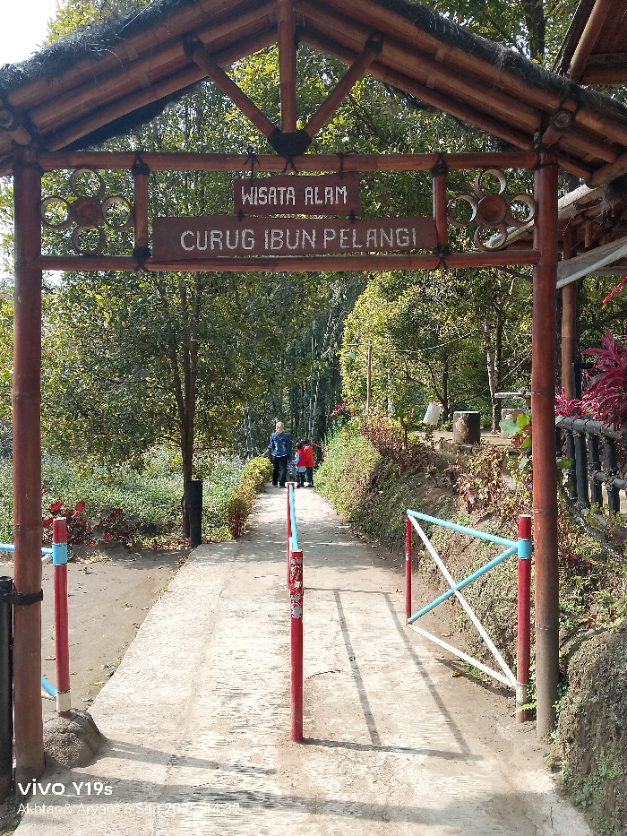 The image captures the entrance to Curug Ibun Pelangi (Rainbow Dew Waterfall), a natural tourist destination located in Sukadana, Indonesia. The foreground features a rustic archway constructed from dark wooden or bamboo-like logs, supporting a thatched roof. Two wooden signs hang from the arch's crossbeam: the upper sign reads "WISATA ALAM" (Natural Tourist Destination), and the lower sign reads "CURUG IBUN PELANGI." A circular, wooden, flower-like decorative element is affixed to the left side of the crossbeam.
A light-colored, possibly concrete, pathway extends from under the archway, leading uphill and curving gently to the left into the distance. Along the pathway, red poles with light blue accents are visible. On the left, two such poles are embedded in the ground, acting as partial barriers. On the right, a low barrier or gate composed of red vertical posts and white and light blue diagonal crossbars is present.
Three individuals are walking away from the camera on the pathway in the mid-ground. The lead person, an adult, is dressed in a light-colored long-sleeved top and dark trousers. Following directly behind are two children, both wearing red tops and dark trousers.
The environment is densely vegetated, with various green trees and shrubs bordering both sides of the pathway. Some reddish-purple foliage is visible near the archway on the right. In the background, to the right of the path, a stone or concrete picnic table and benches are partially visible amidst the greenery. The scene is illuminated by bright natural sunlight, casting distinct shadows from the entrance structure and poles onto the pathway.